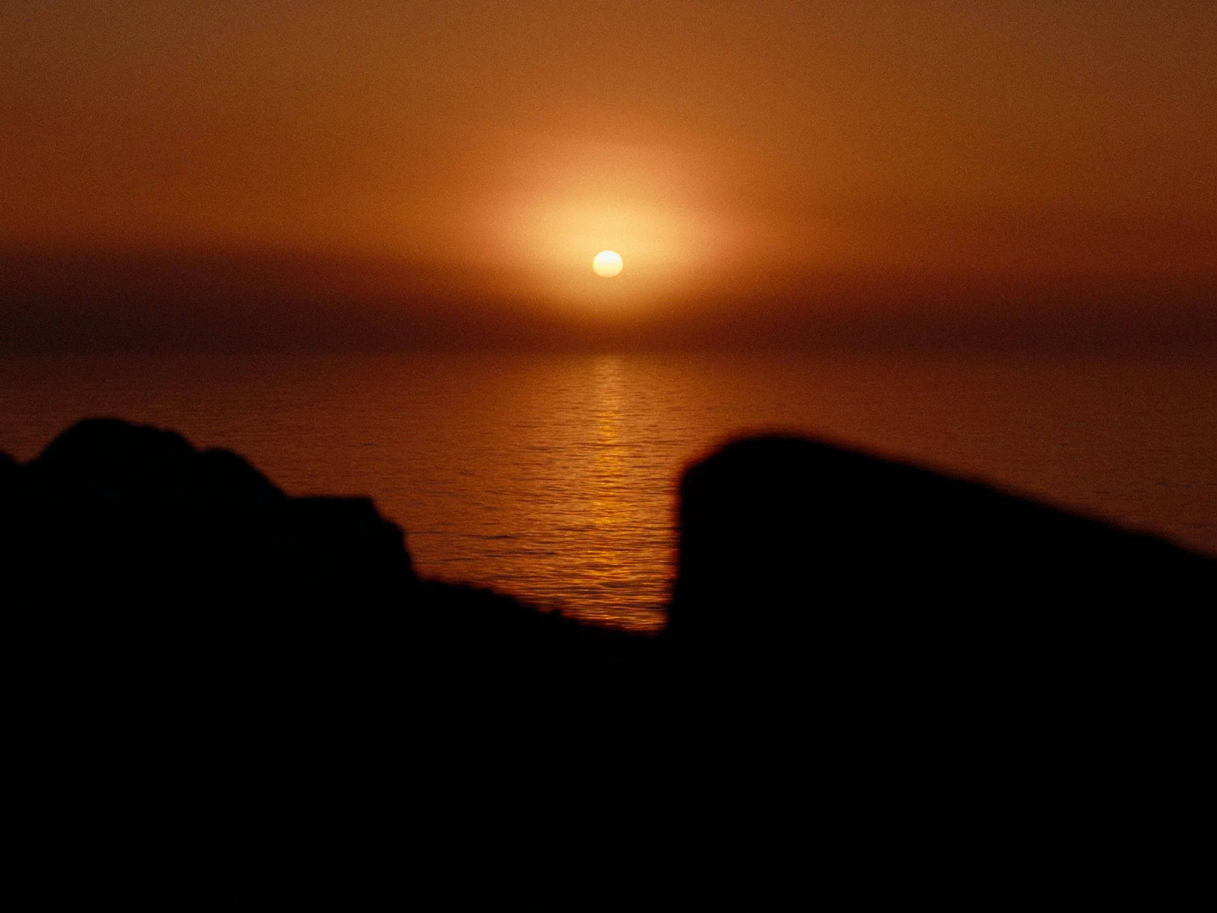 A sunset over the ocean with the sun partially visible near the horizon, with dark silhouettes of rocks or land in the foreground.