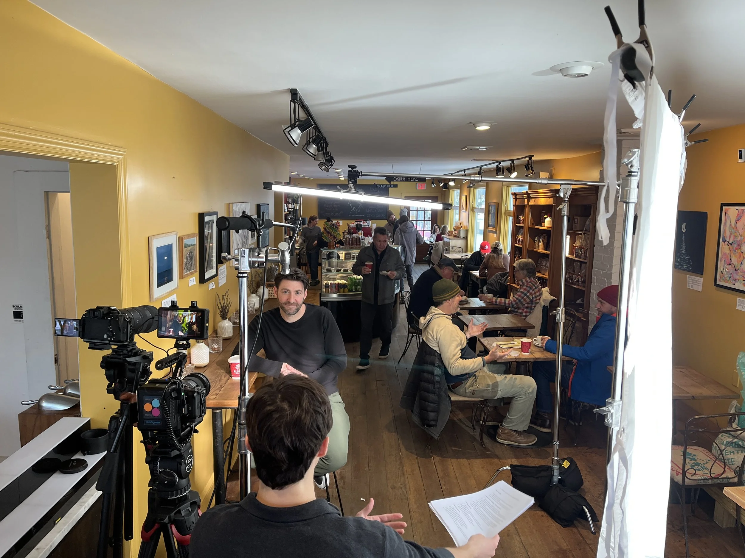 Interior of a busy cafe with people sitting at tables, some reading and others holding drinks, a person filming with professional equipment, and yellow walls decorated with framed artwork.