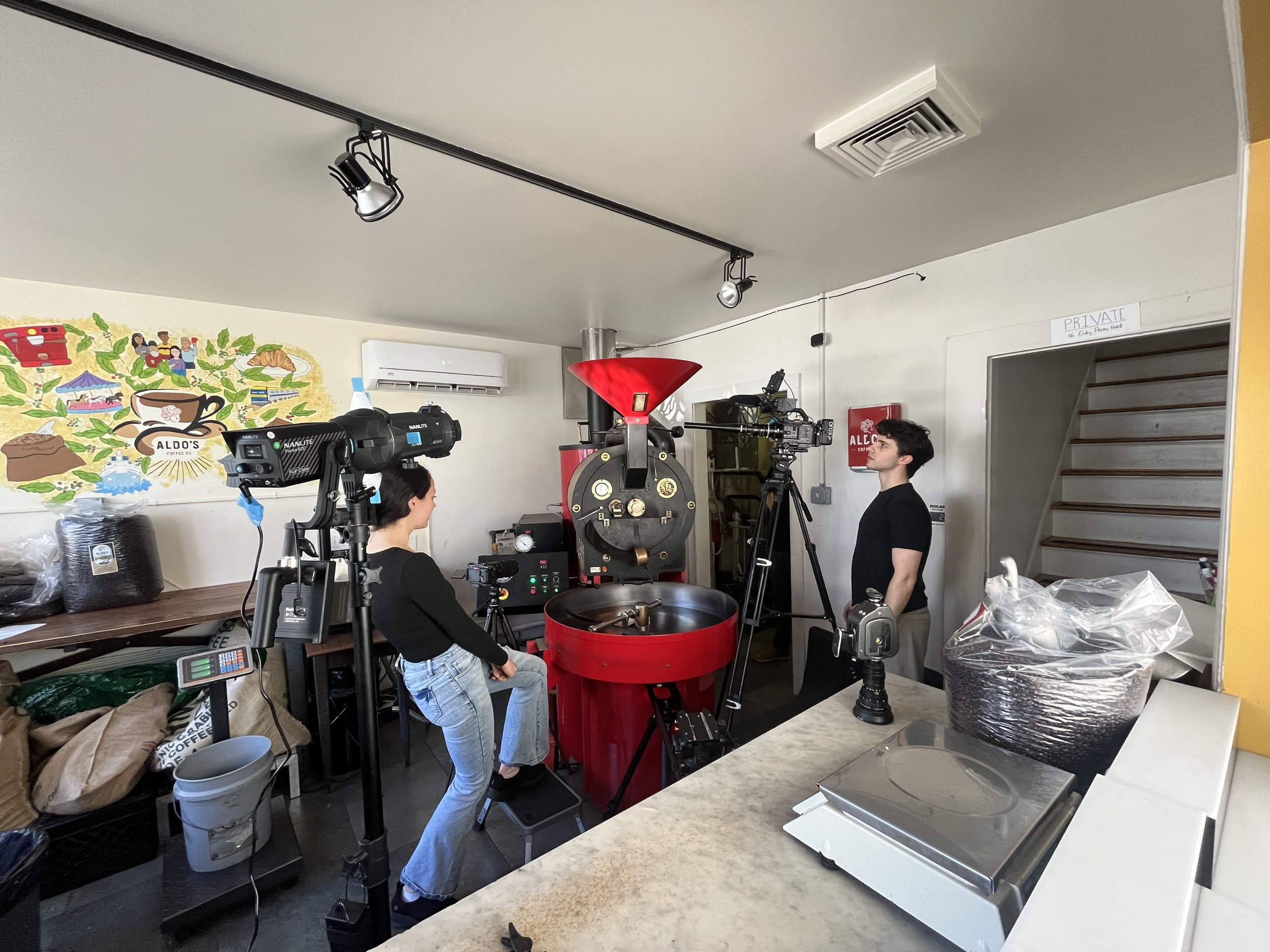 A coffee shop interior with people working with cameras and equipment, a colorful mural on the wall, coffee supplies on the counter, and a staircase in the background.
