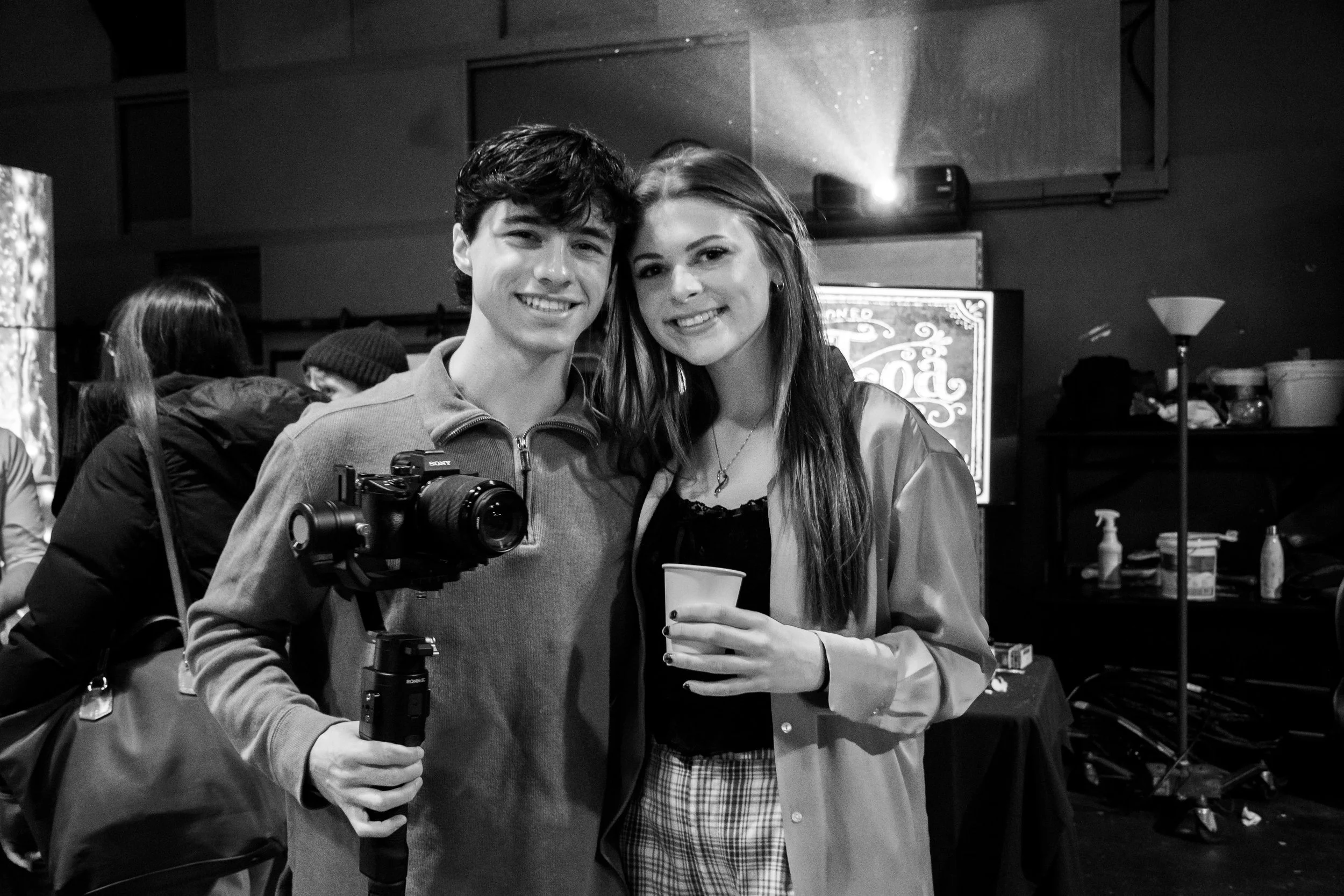 Two young women posing together at an event, one holding a camera on a tripod and the other holding a drink, in a crowded indoor setting.