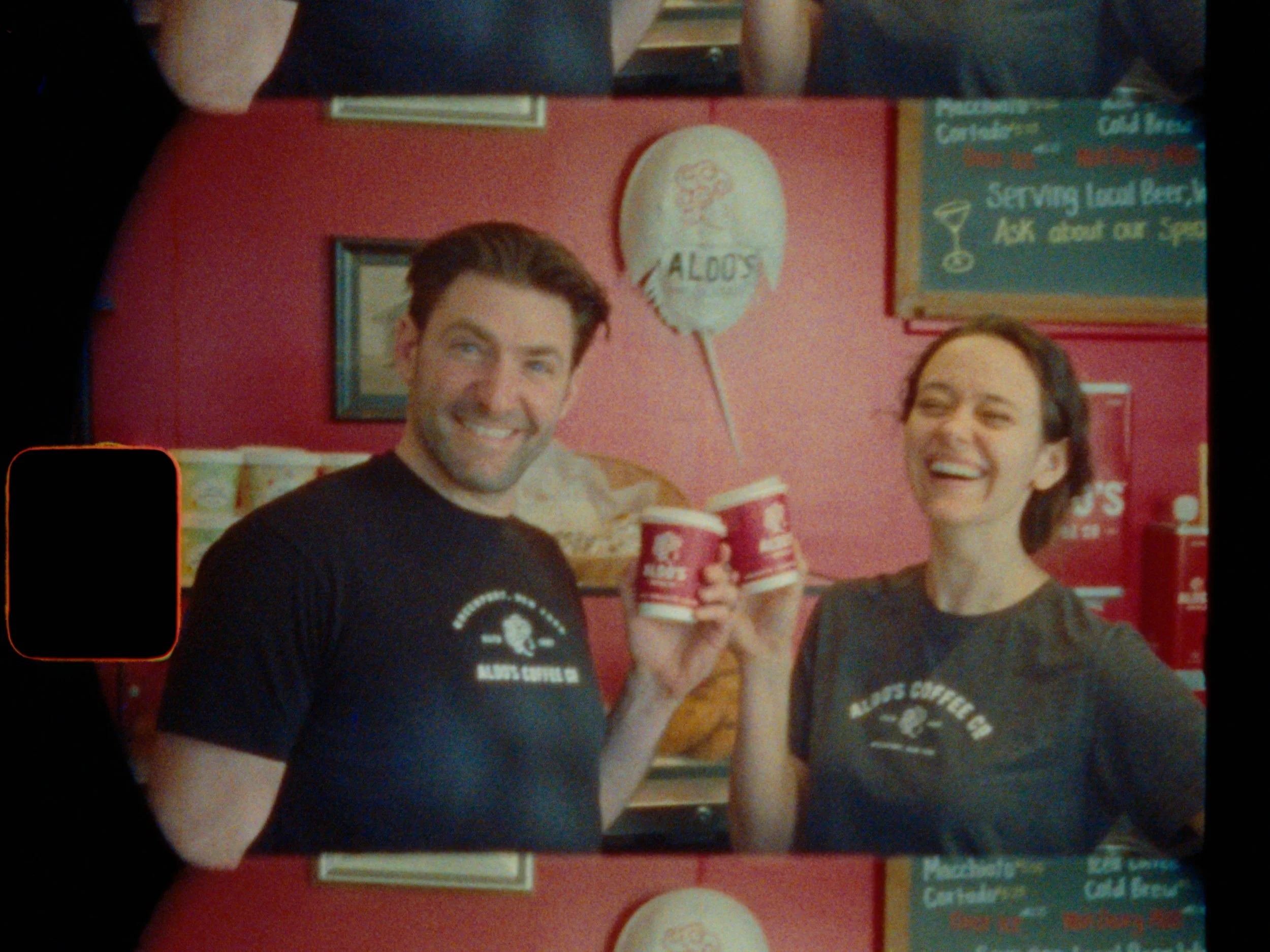 Two people smiling and holding cups of coffee inside a fast food restaurant, with a red wall and menu boards in the background.