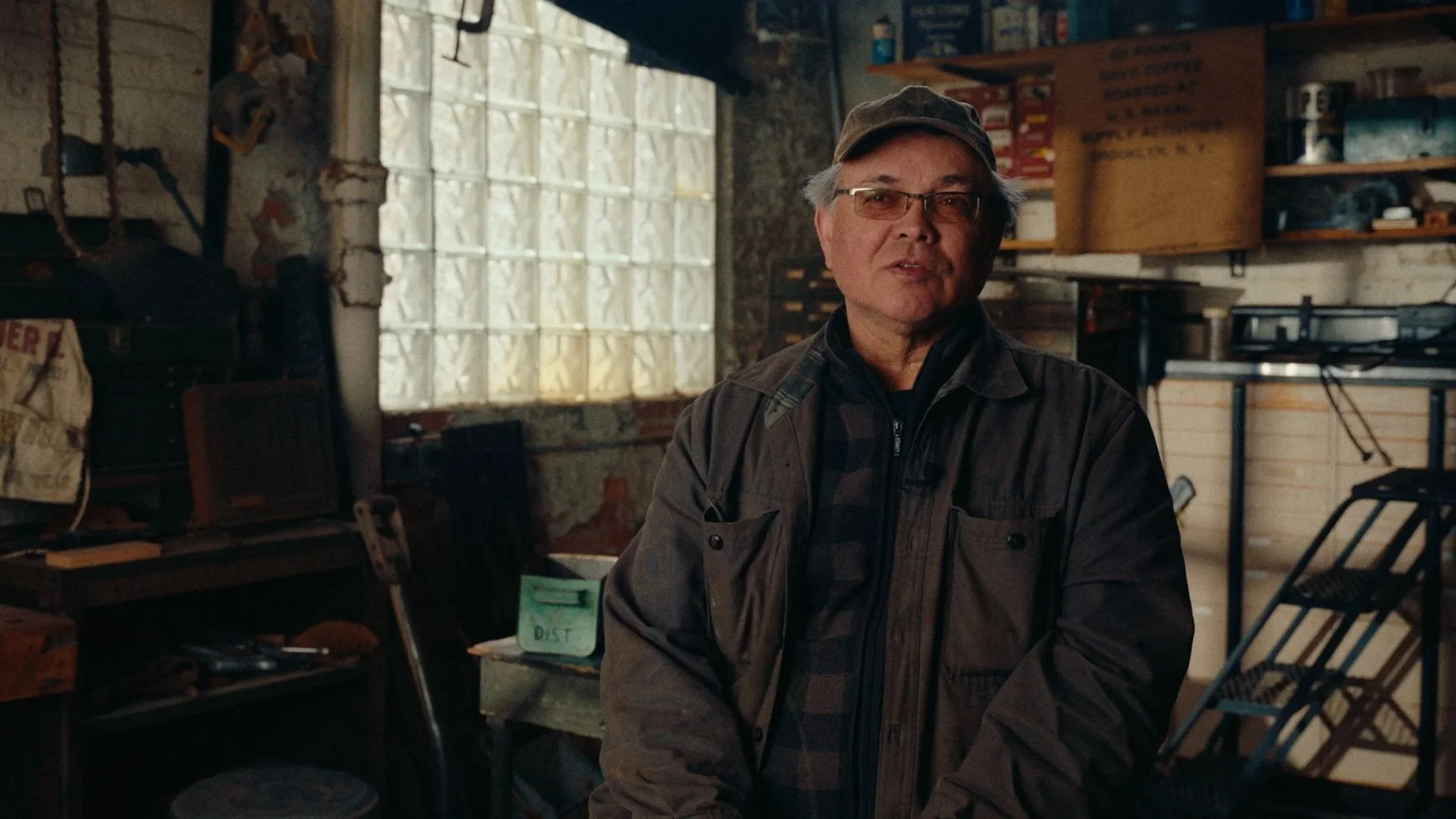 An elderly man wearing glasses and a cap standing inside a cluttered workshop or garage, with shelves filled with boxes and tools in the background.
