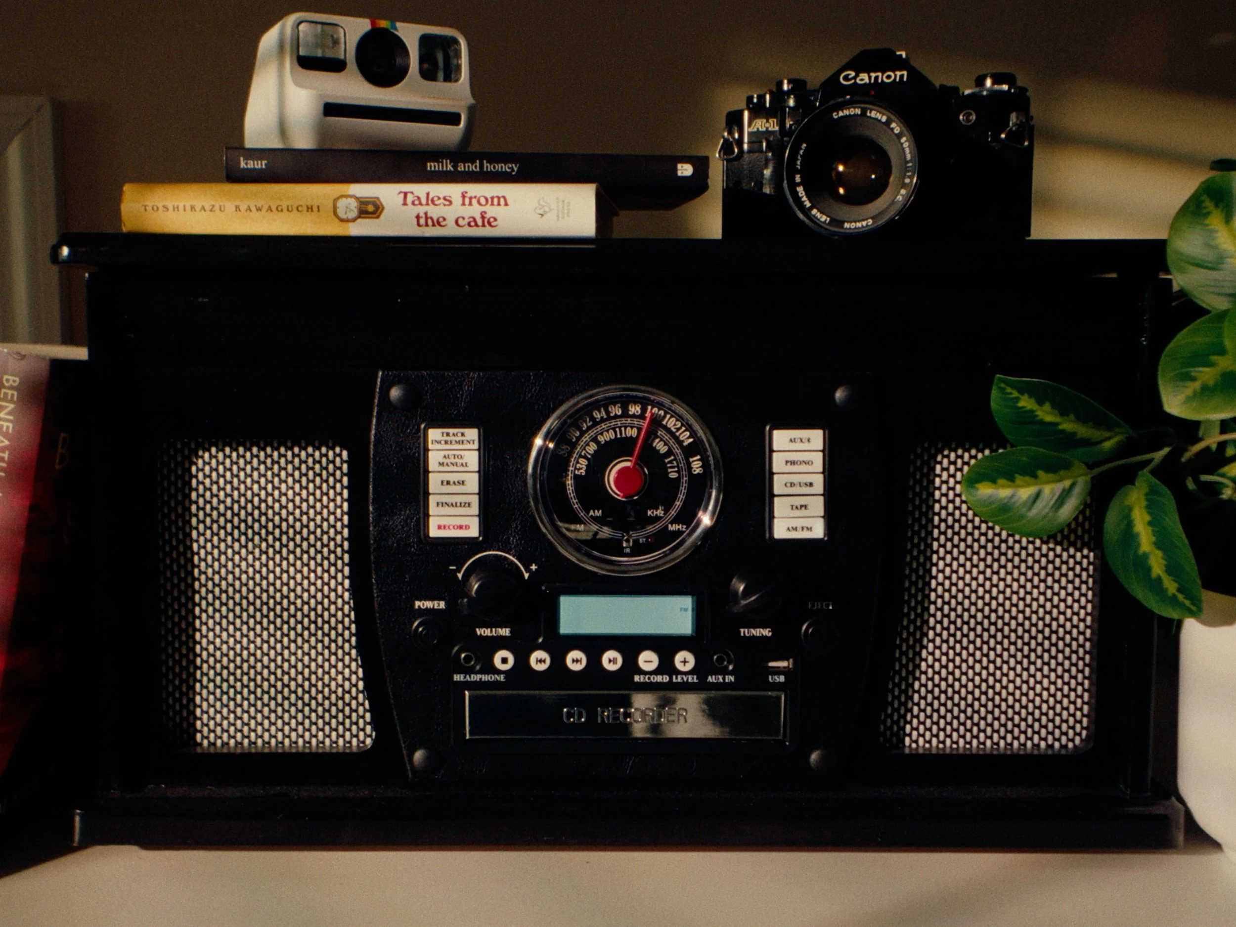 A black vintage radio with a round dial and buttons, topped with two books titled 'milk and honey' and 'Tales from the cafe,' a camera, and a small white robot.