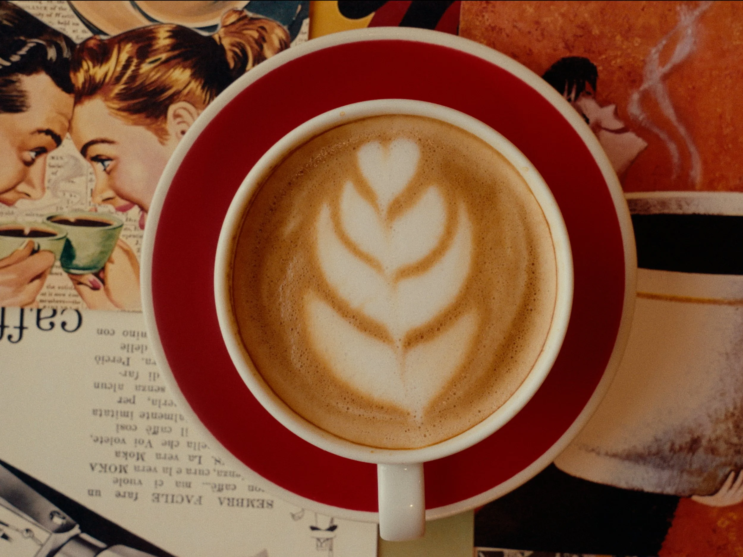 Close-up of a latte in a white mug with a red interior, featuring latte art in the shape of a tulip. The mug is on a table next to a magazine or newspaper with pictures of people holding cups of coffee.