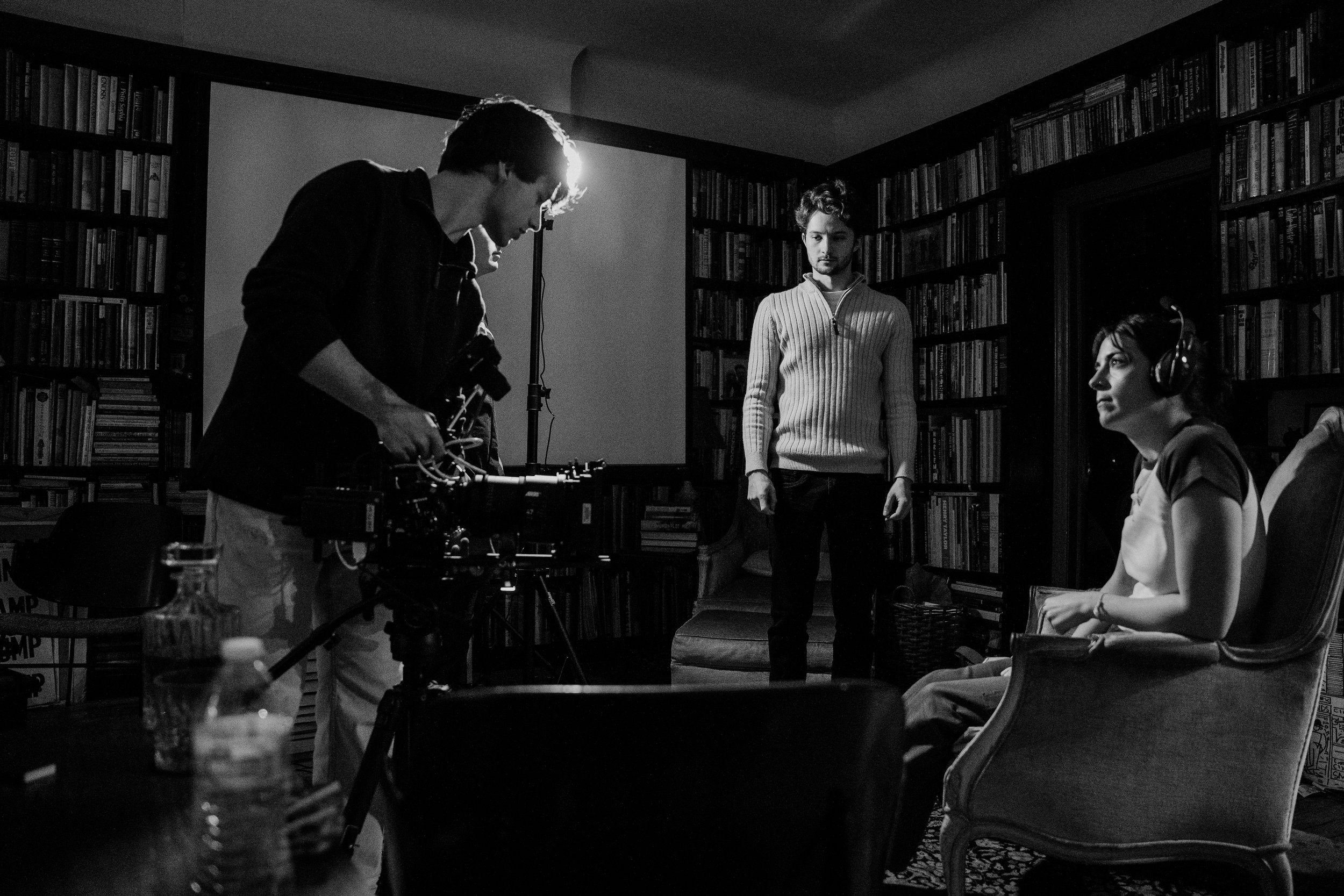 Black and white photo of a film set or interview room with three people: a man on the left adjusting camera equipment, a woman on the right sitting in a chair wearing headphones, and a man standing in the middle watching. The room has bookshelves filled with books.