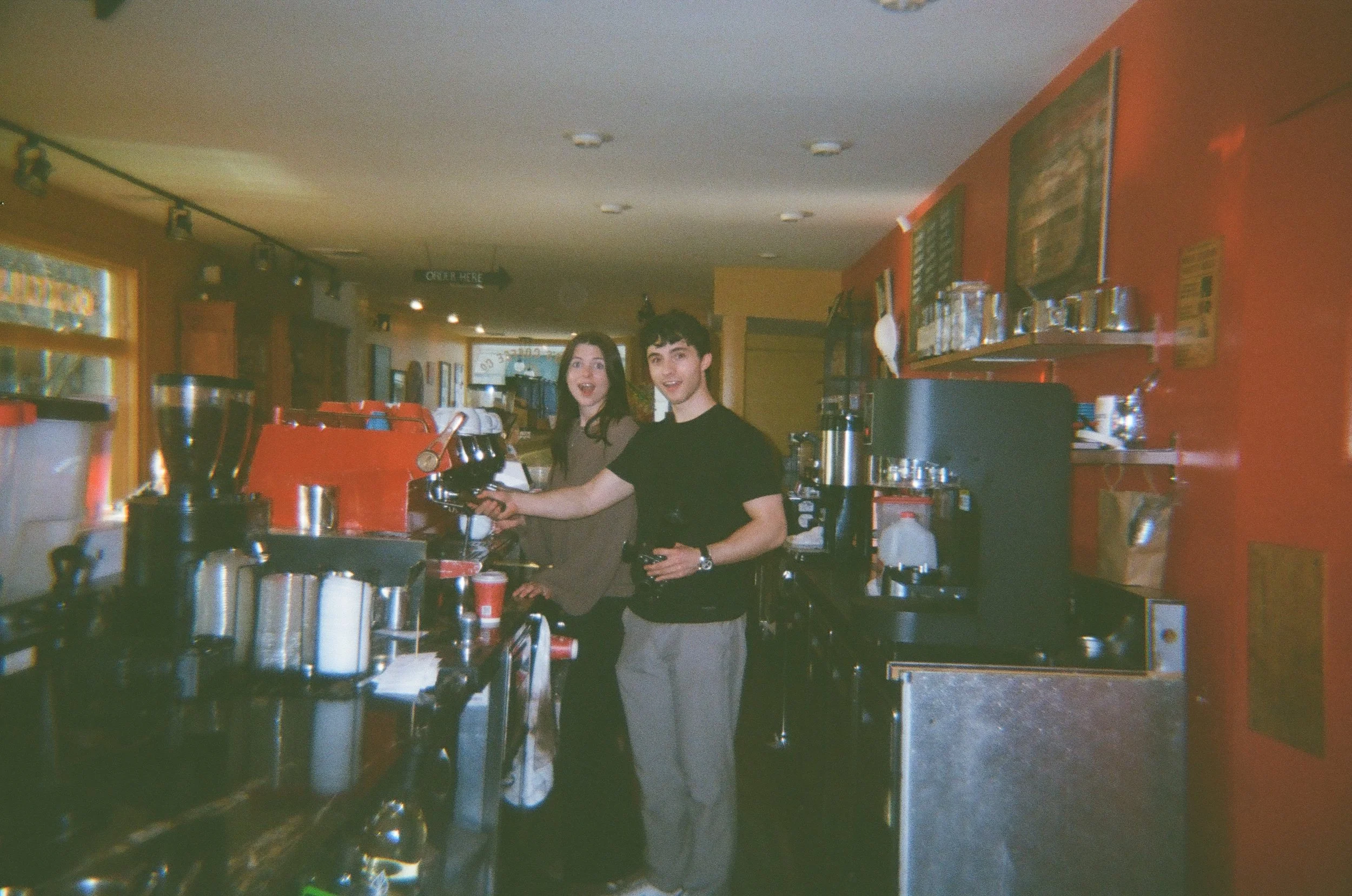 Two young people, a woman and a man, working behind a coffee bar in a cafe, smiling at the camera.