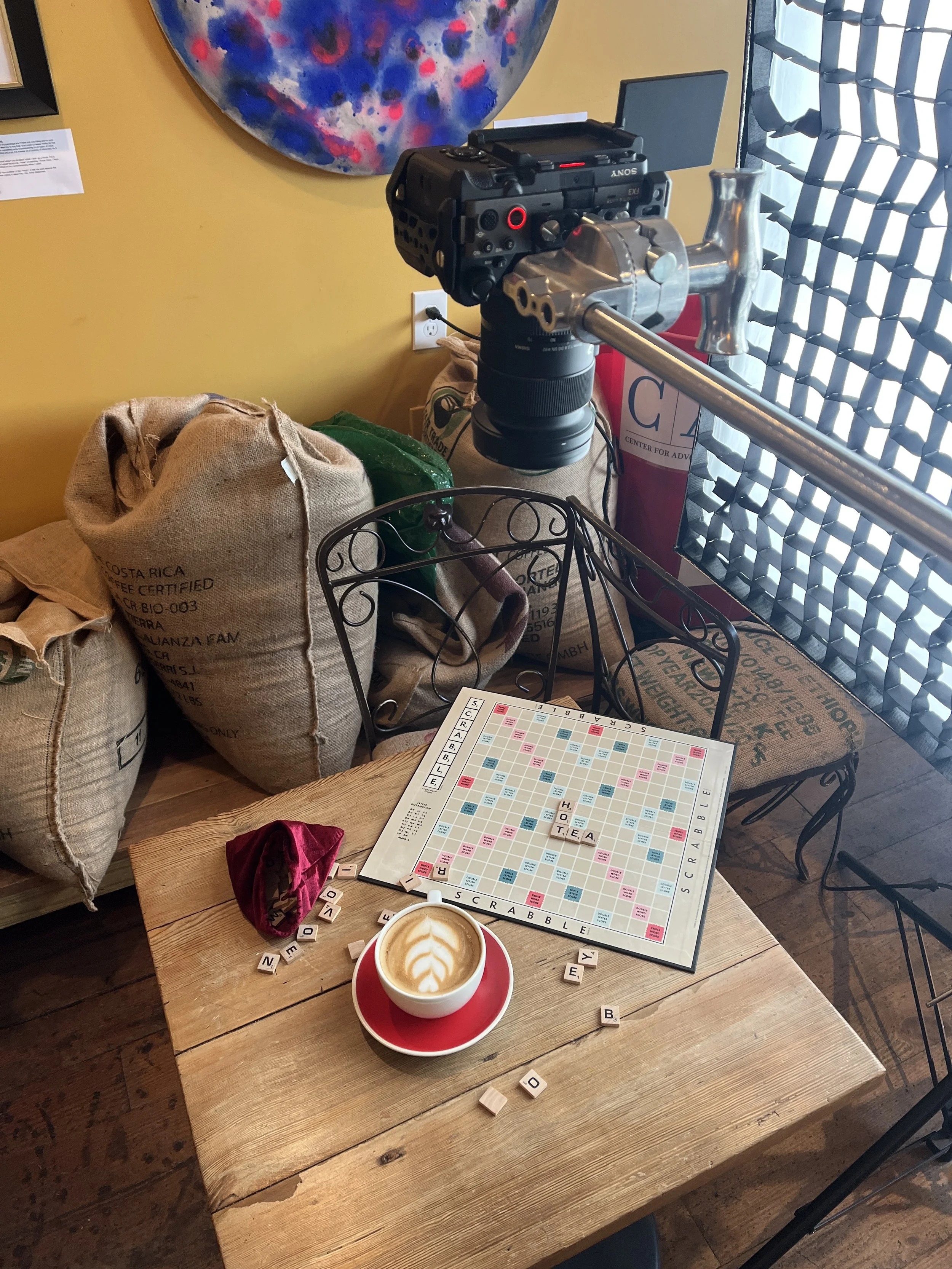 A cozy indoor scene featuring a Scrabble board with words like 'HOT' and 'TEA', wooden letter tiles, a cup of coffee with latte art, and various bags and containers on a wooden table. A camera is mounted on a tripod pointing towards the scene, with a colorful abstract artwork on the yellow wall behind.