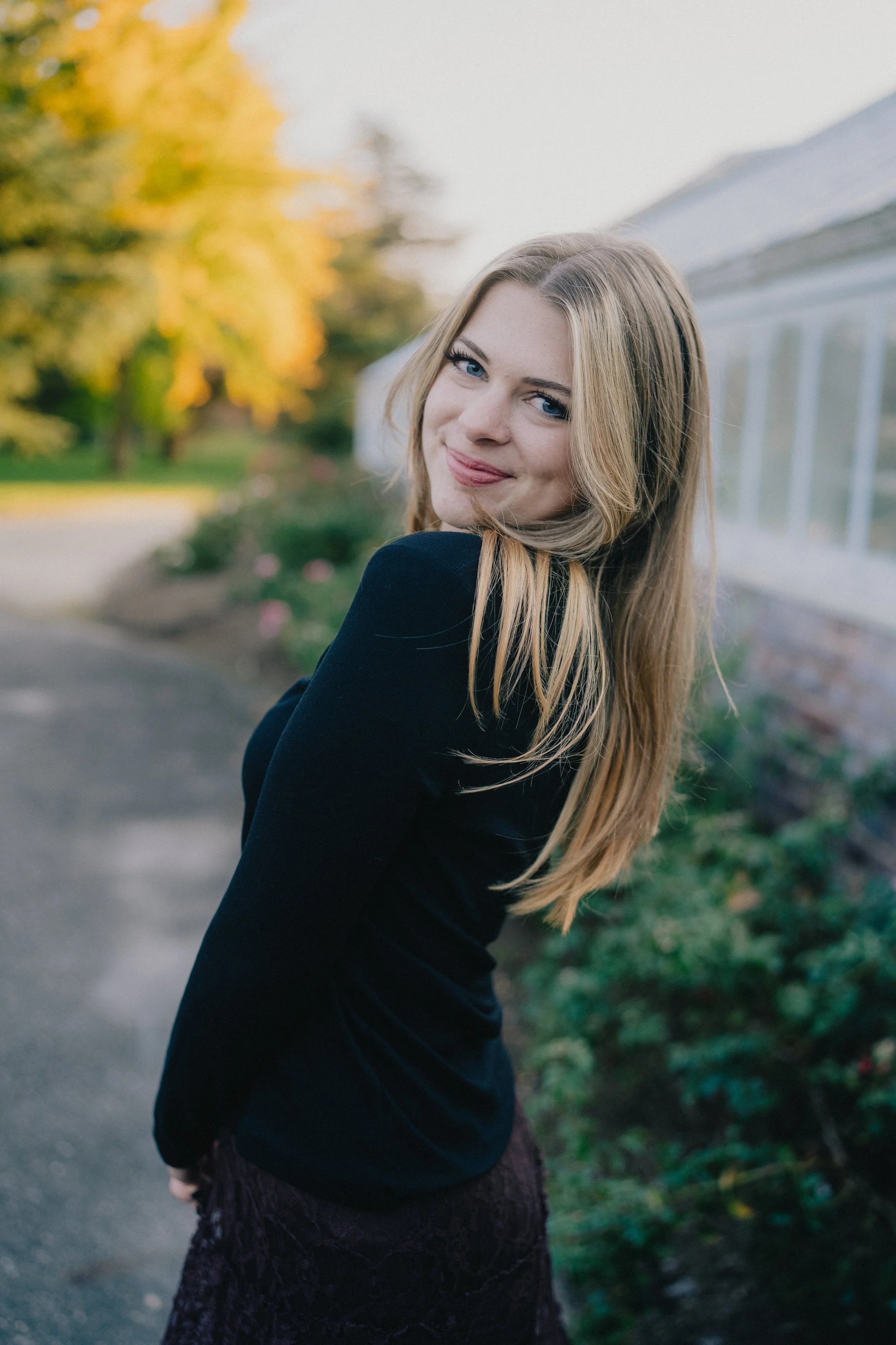 A young woman with long blonde hair and blue eyes is smiling at the camera, standing outdoors with trees and a greenhouse in the background.