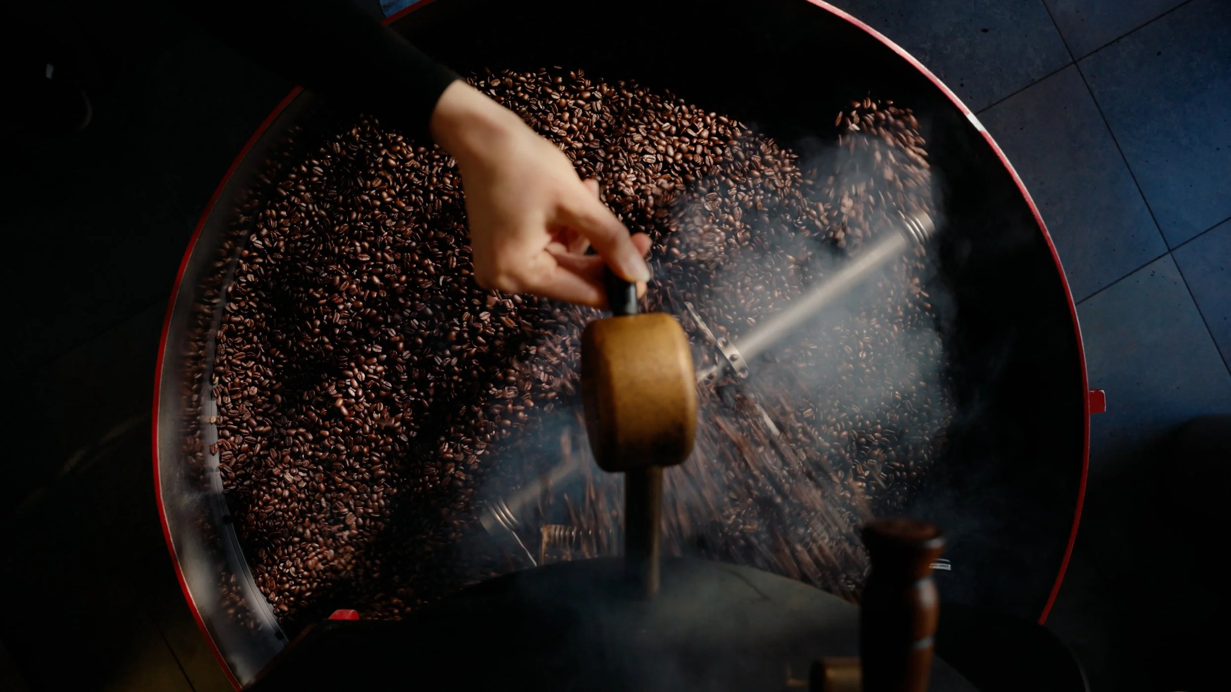 Close-up of roasted coffee beans in a large metal drum, with a person stirring or raking the beans, creating steam and smoke.