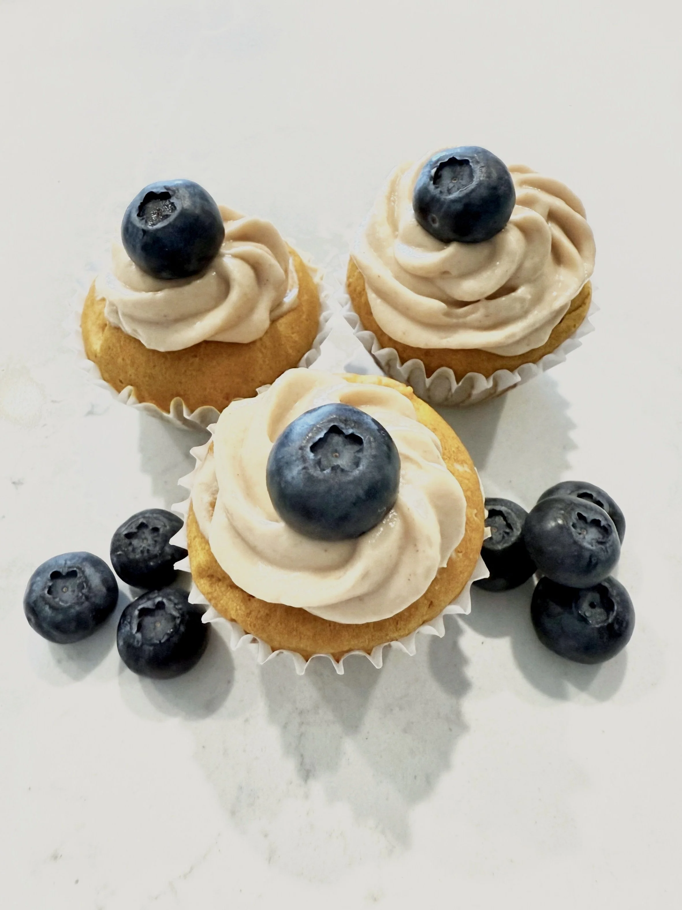 Three cupcakes with blueberry toppings and several loose blueberries on a white surface.