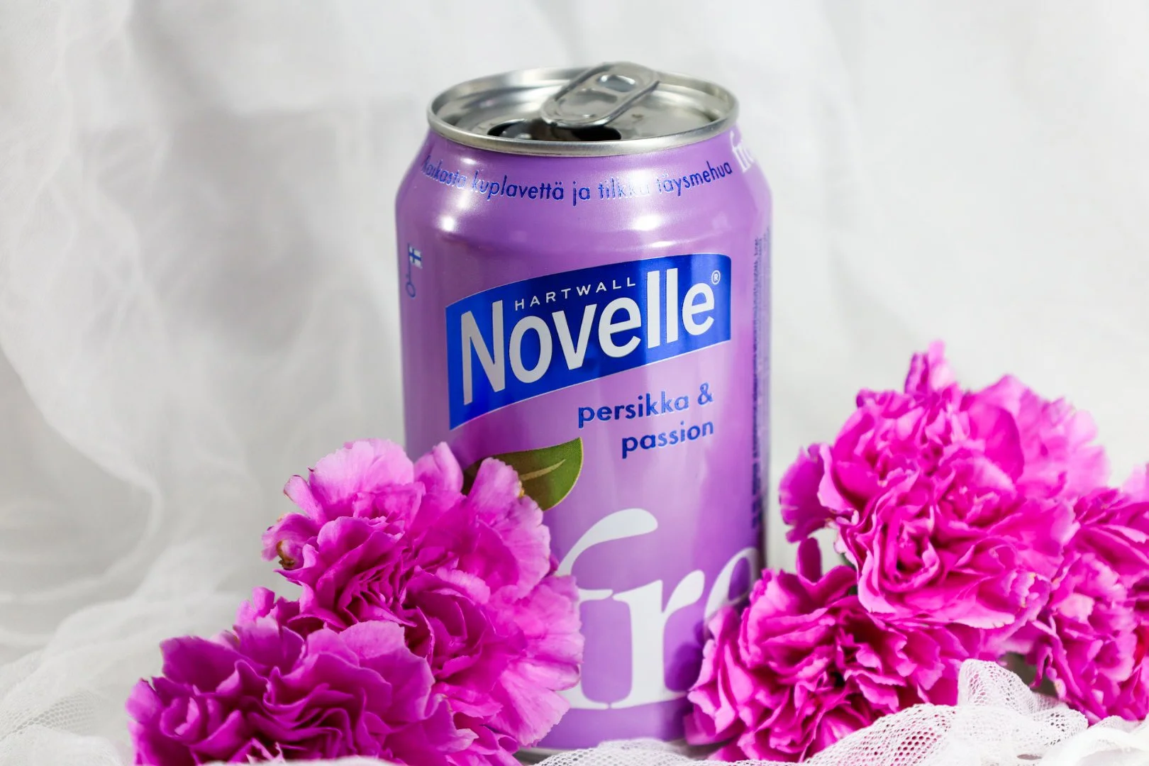 Violetti Novelle-persikkainen ja passionhedelmäinen juomatölkki, ympärillä vaaleanpunaisia neilikoita ja valkoinen kangas taustalla.