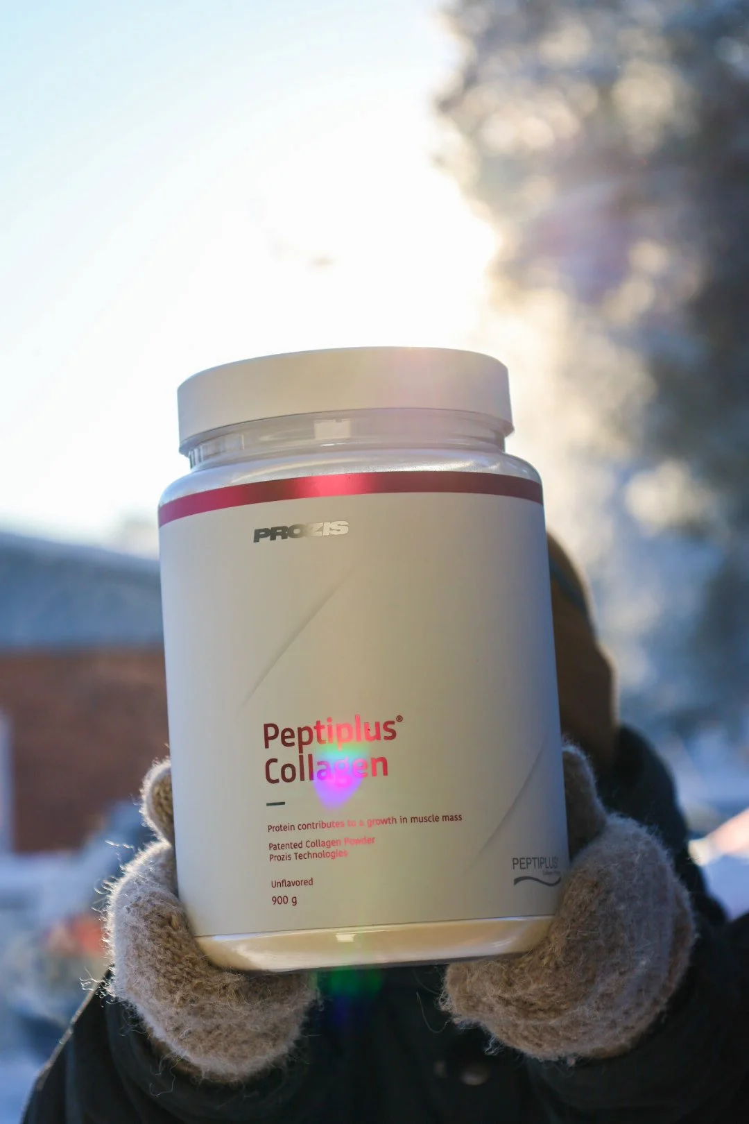 Käsineillä pideltävä valkoinen ravintolisäpurkki sisältää Peptiplus Collagen -tuotetta, taustalla kirkas auringonvalo ja talvinen maisema.