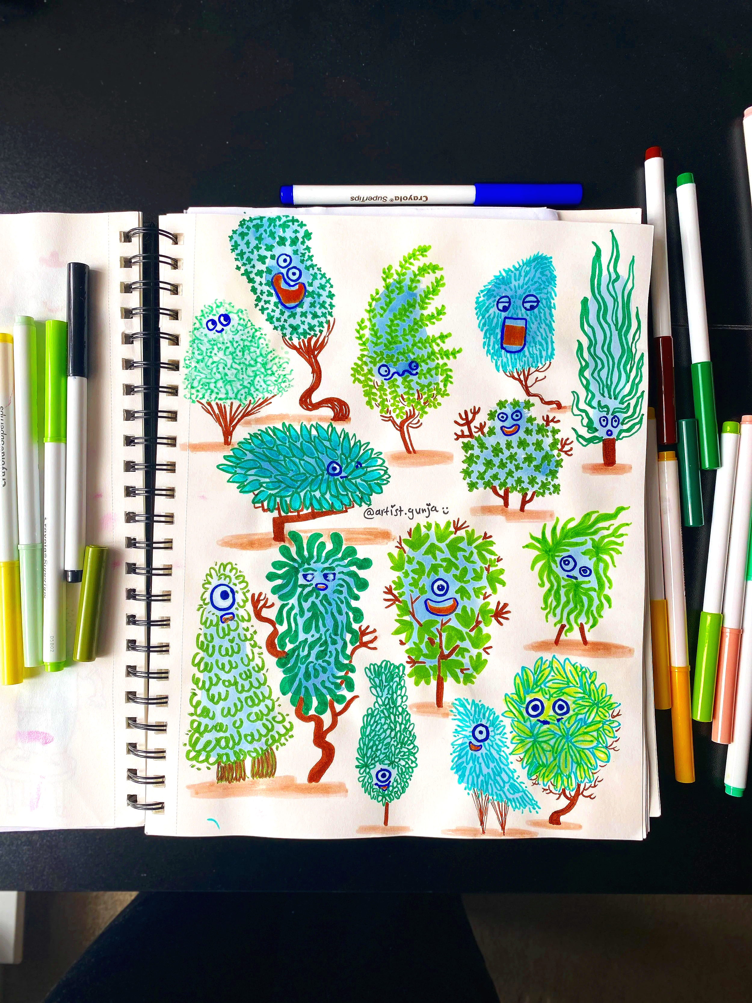 Colorful illustrated trees with faces, drawn with markers on a sketchbook, surrounded by various colored markers.