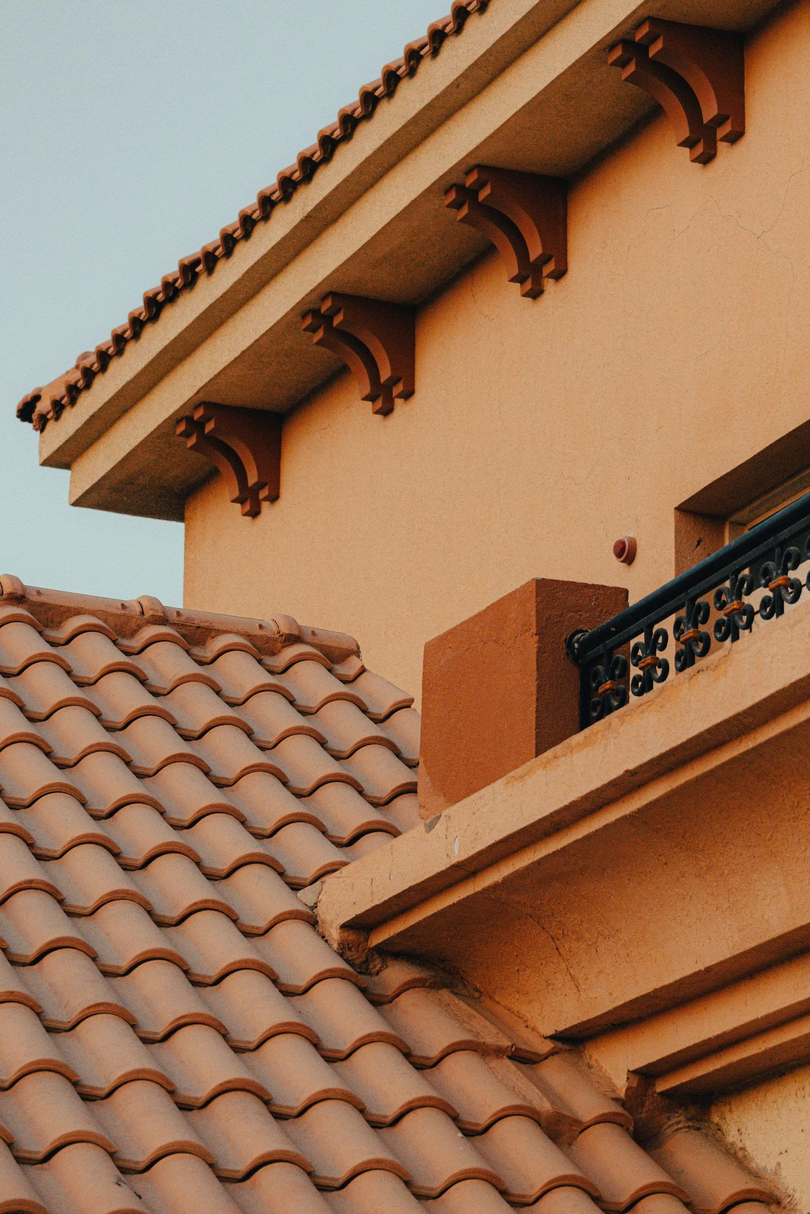 Close-up of a building with terracotta roof tiles, stucco walls, decorative corbels under the eaves, and a small balcony with a black wrought-iron railing.