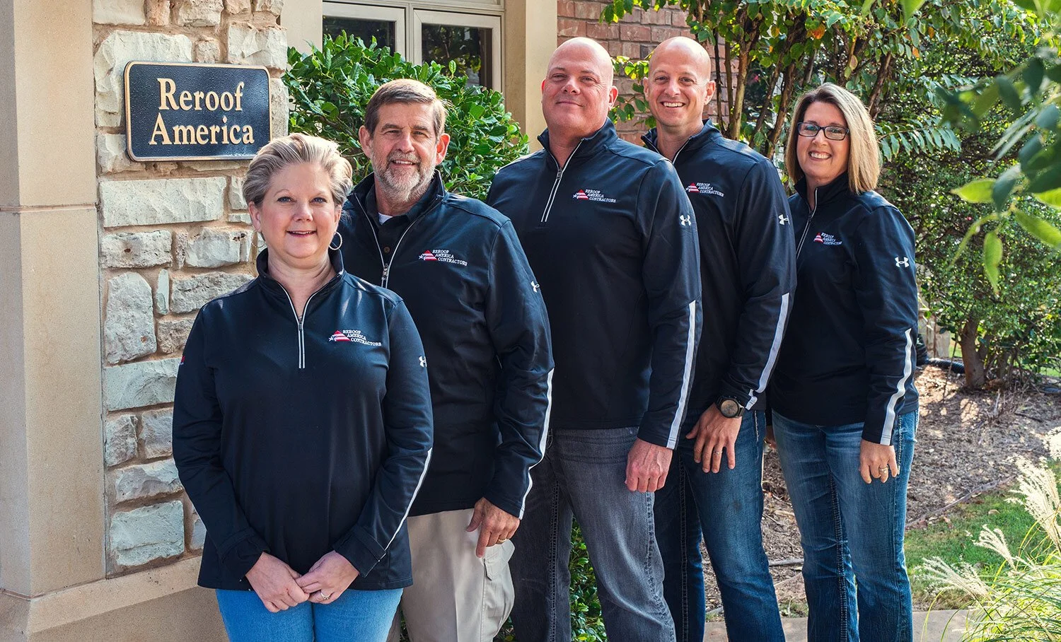 Group of five smiling people wearing matching dark jackets standing outdoors in front of a house with a sign that reads 'Reroof America'.
