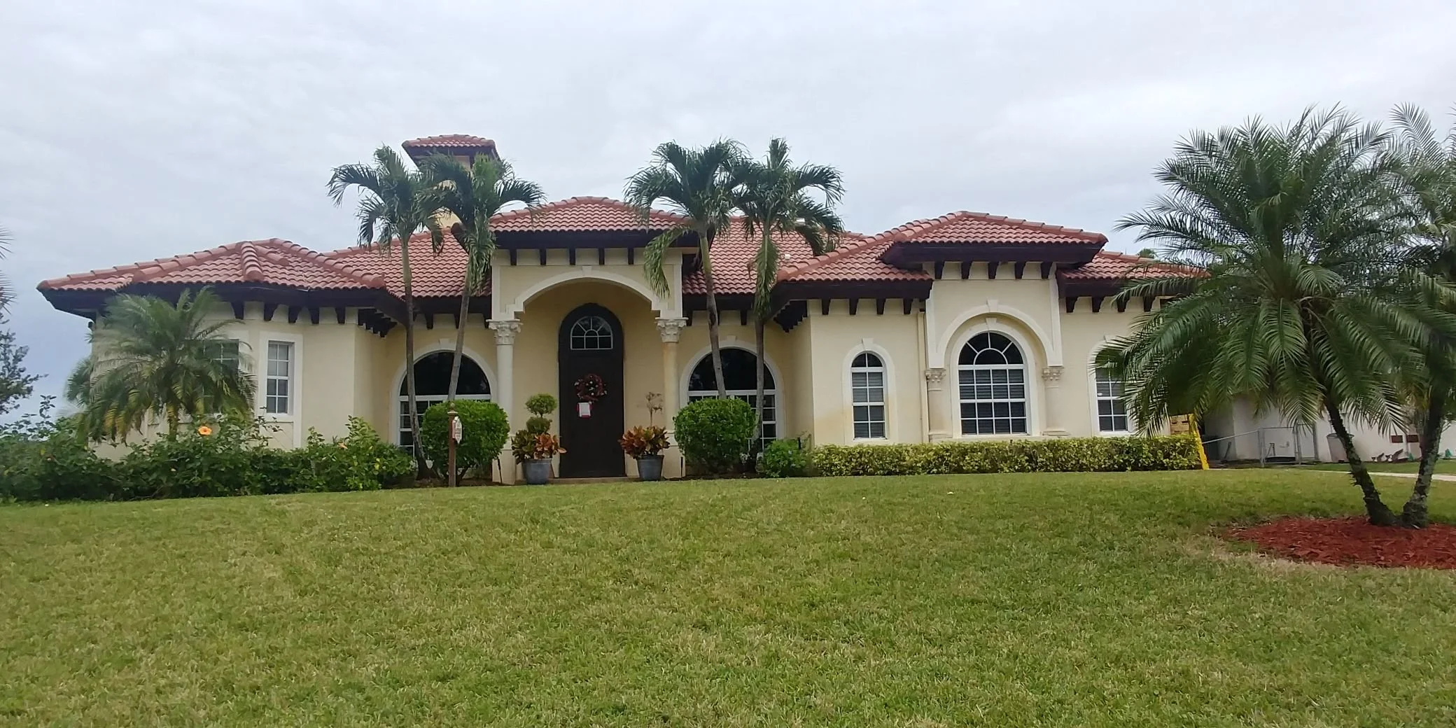 Large Mediterranean-style house with a red-tile roof, arched windows, and a well-maintained lawn with palm trees and shrubs.