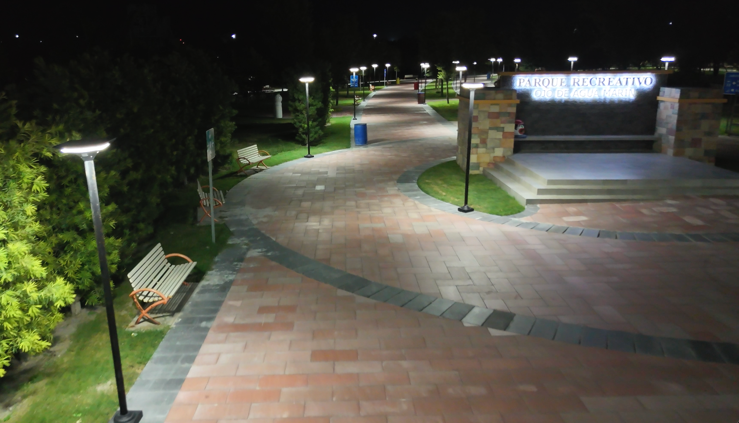 Camino peatonal en un parque recreativo con bancos, lámparas y áreas verdes, entrada con cartel que dice 'Parque Recreativo Ojo de Agua Marin' durante la noche.