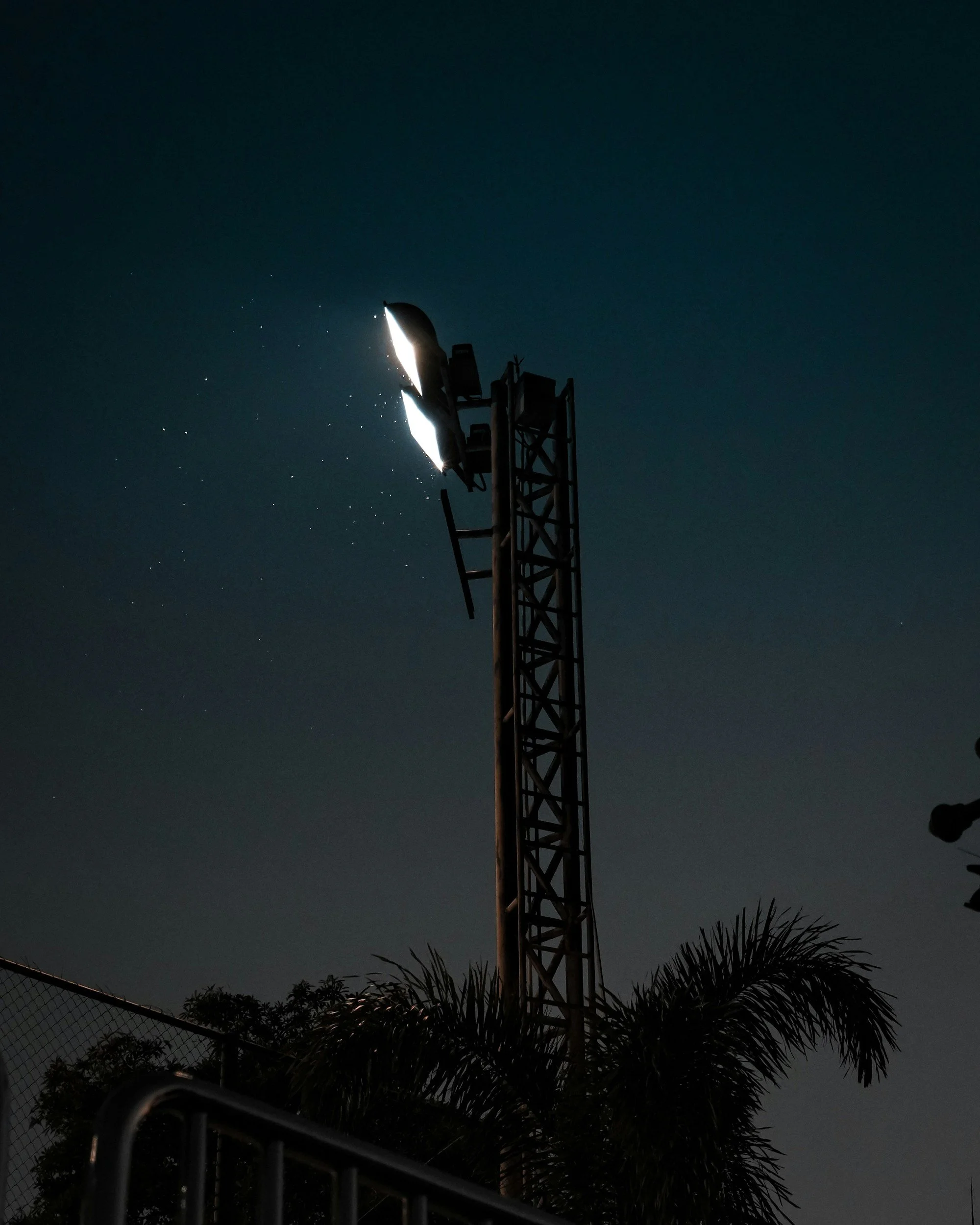 Una torre de telecomunicaciones grande contra un cielo nocturno, con estrellas visibles y palmeras en primer plano.