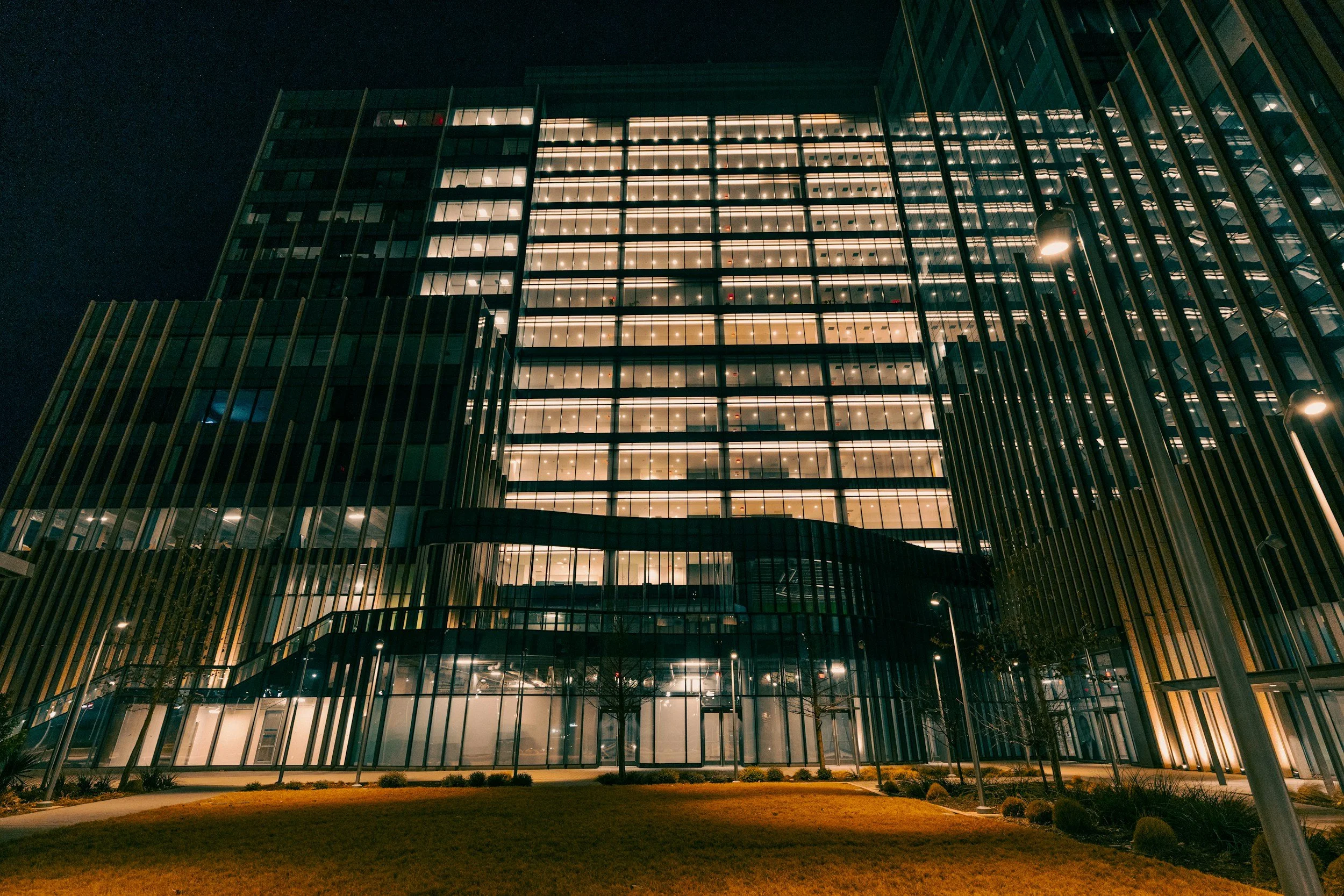 Edificio moderno de varias plantas con cristaleras, iluminado en la noche, con área de césped y luminarias en la parte inferior.