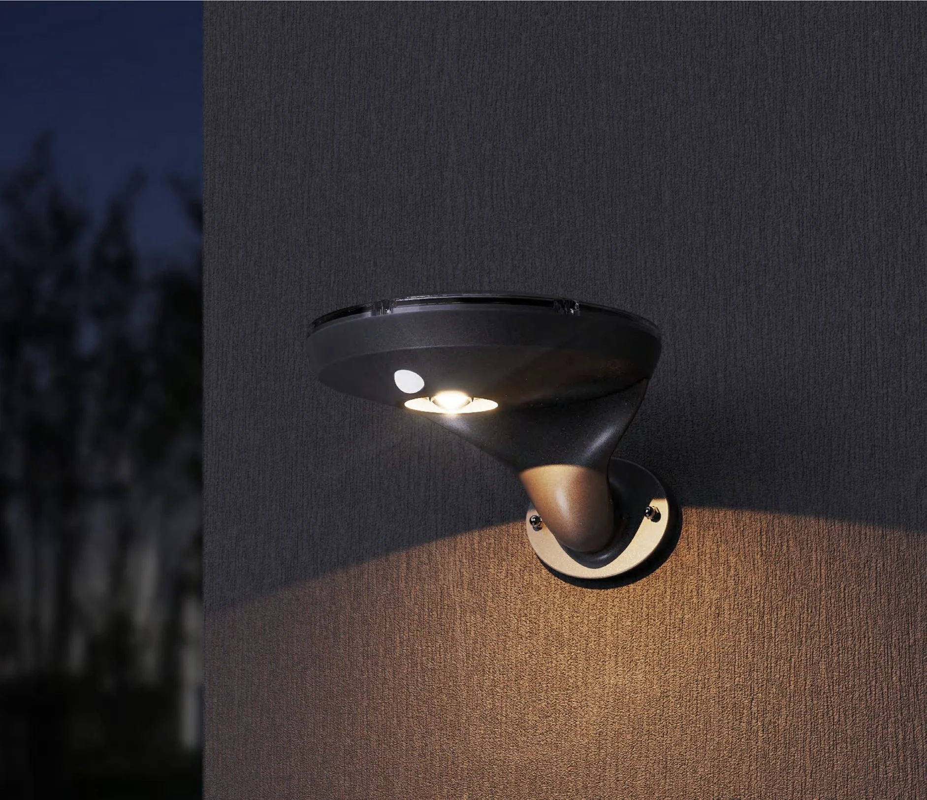 Lámpara de pared negra iluminando una superficie exterior, con un fondo oscuro de árboles desenfocados.