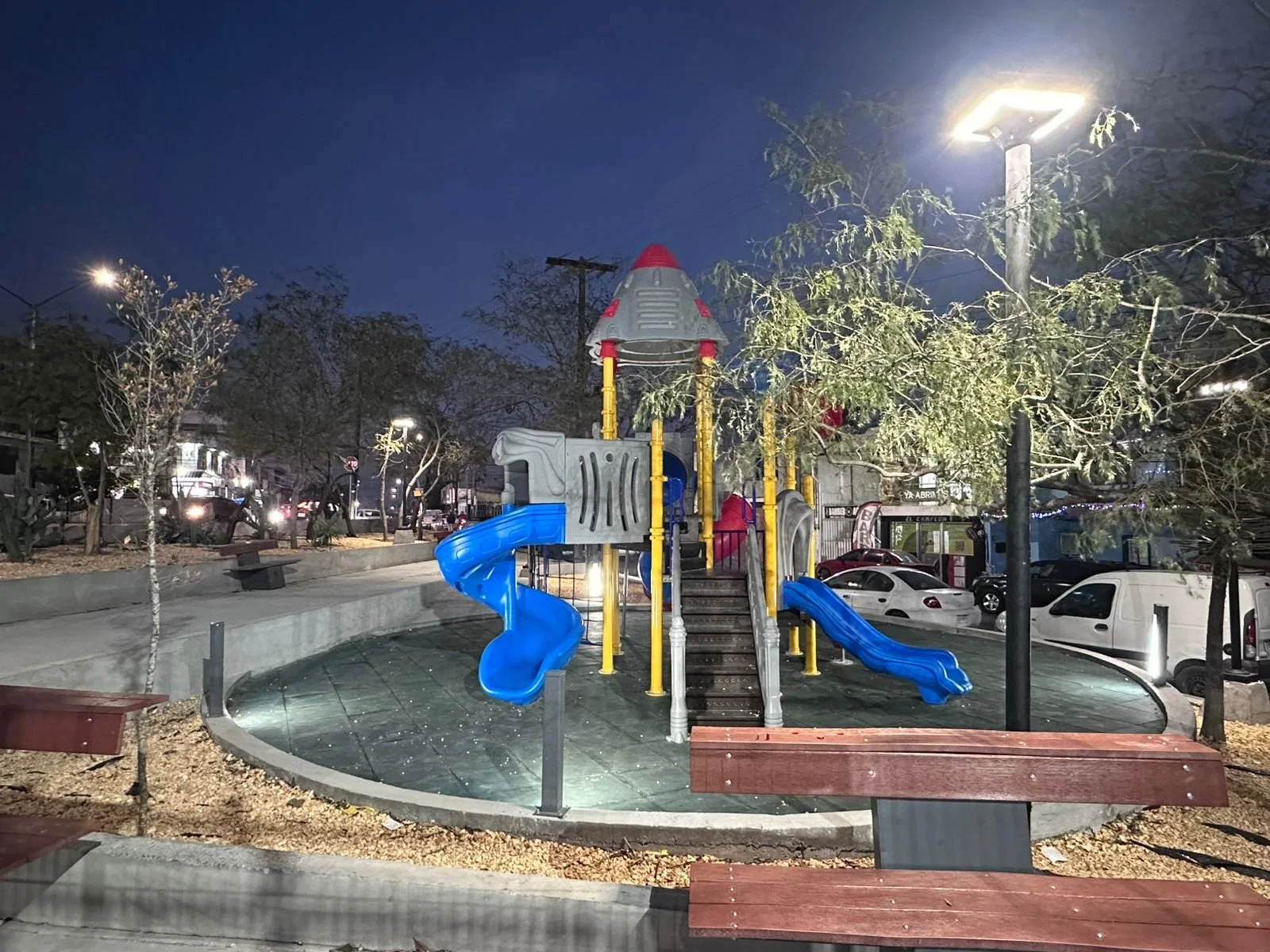 Juegos infantiles en un parque al anochecer, con resbaladilla azul y estructura decorada como cohete, rodeados de árboles con ramas secas y luces en el cielo.