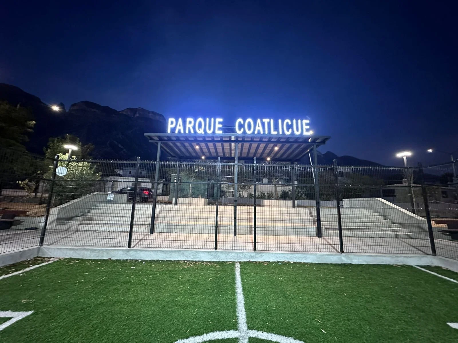 Entrada de un parque llamado 'Parque Coatlucie' con iluminación nocturna, vista desde un campo de césped artificial de fútbol, con montañas al fondo y valla metálica en primer plano.