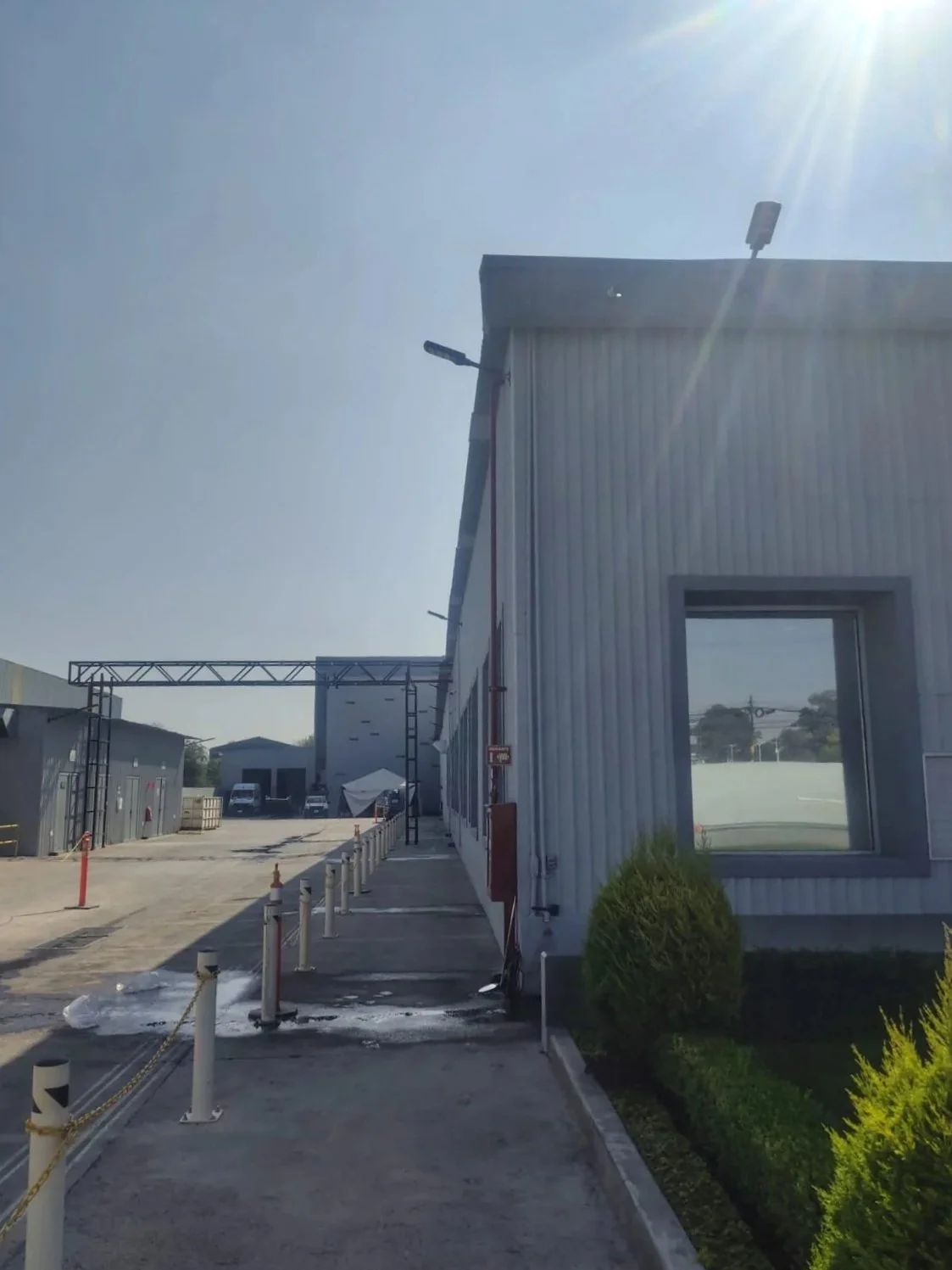 Vista del exterior de un edificio industrial con paredes de metal, una ventana y un camino señalizado con postes blancos y cadenas, con un cielo despejado y sol brillante.