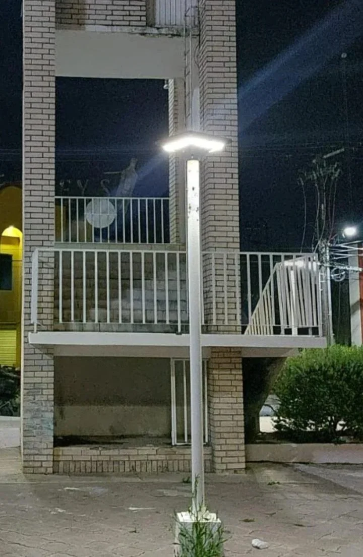 Una farola encendida en la calle frente a un edificio de dos pisos con barandales blancos y paredes de ladrillo. En la planta superior hay una figura de un sacerdote o monje con hábito, visible a través de una ventana.