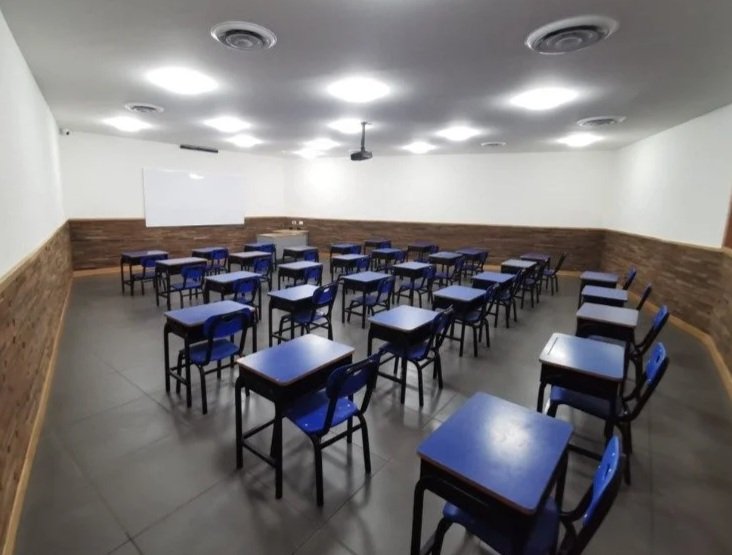 Aula vacía con escritorios y sillas azules, paredes blancas con moldura de madera, y pizarra blanca en la pared frontal.