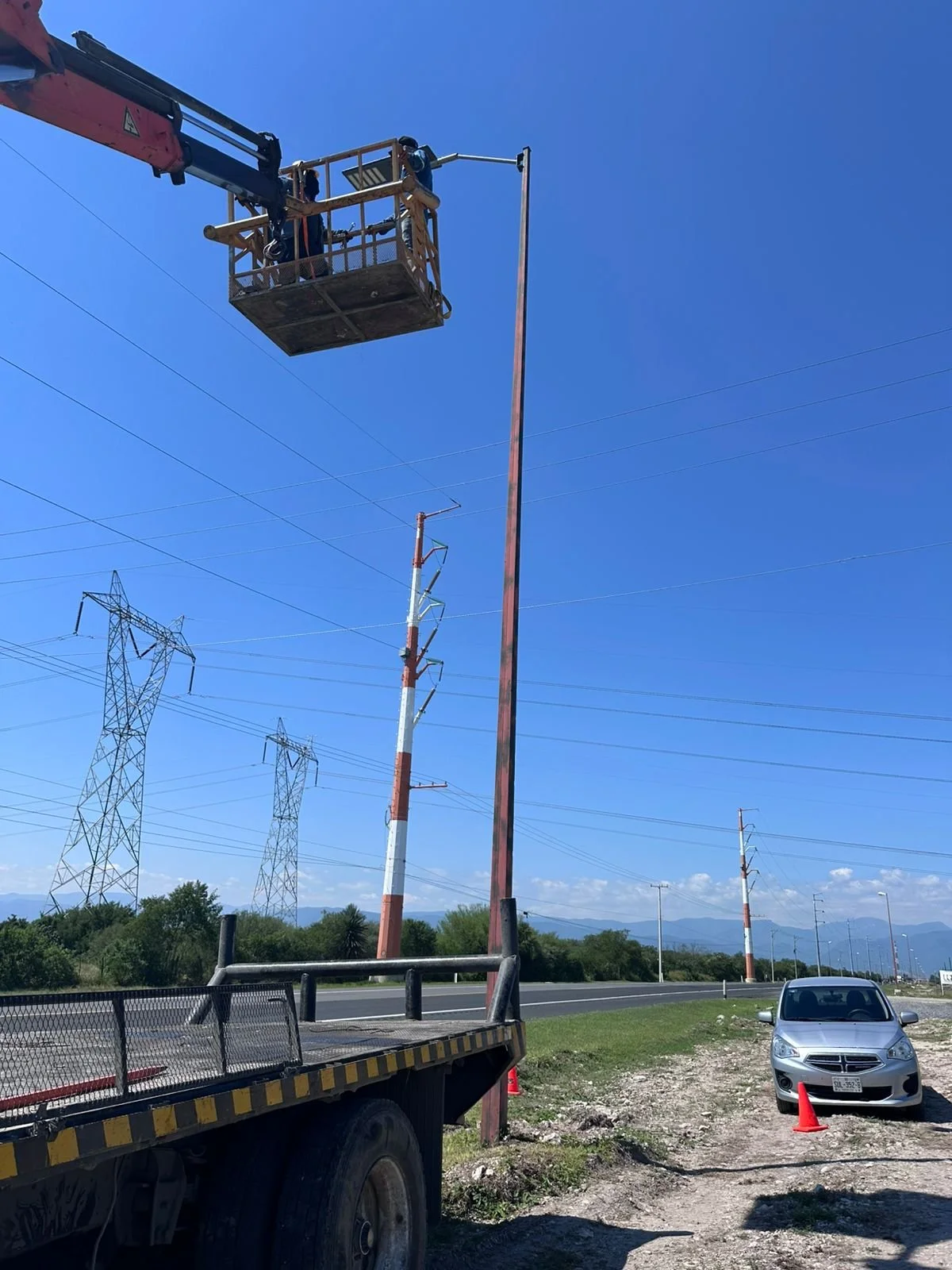 Trabajadores en una plataforma elevadora instalando o reparando postes de electricidad junto a una autopista. Hay un camión de servicios y un auto estacionado con un cono naranja cerca.