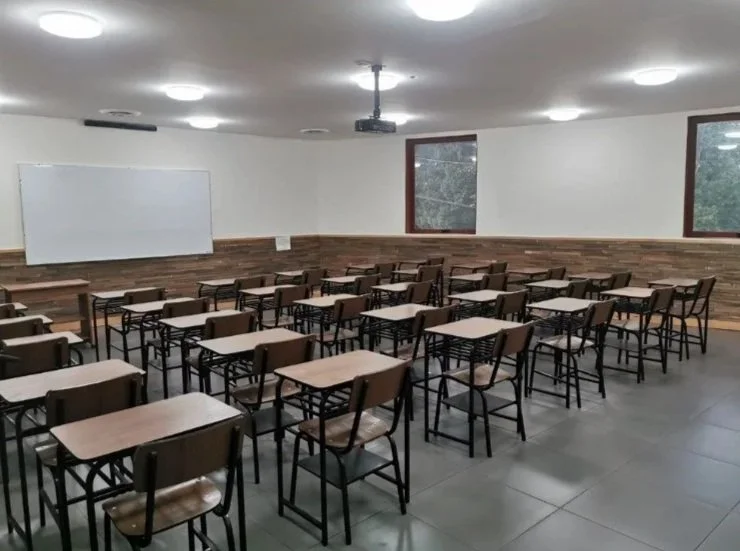 Sala de clases vacía con escritorios y sillas, pizarra blanca, ventanas y techo con lámparas