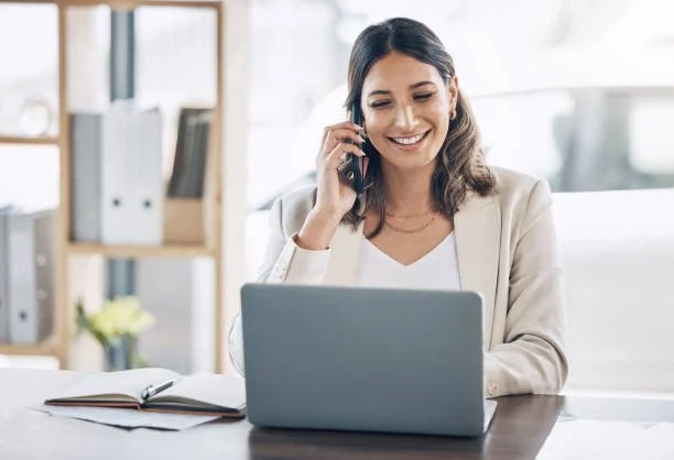 Woman smiling and talking on the phone while working on a laptop at a desk in an office.