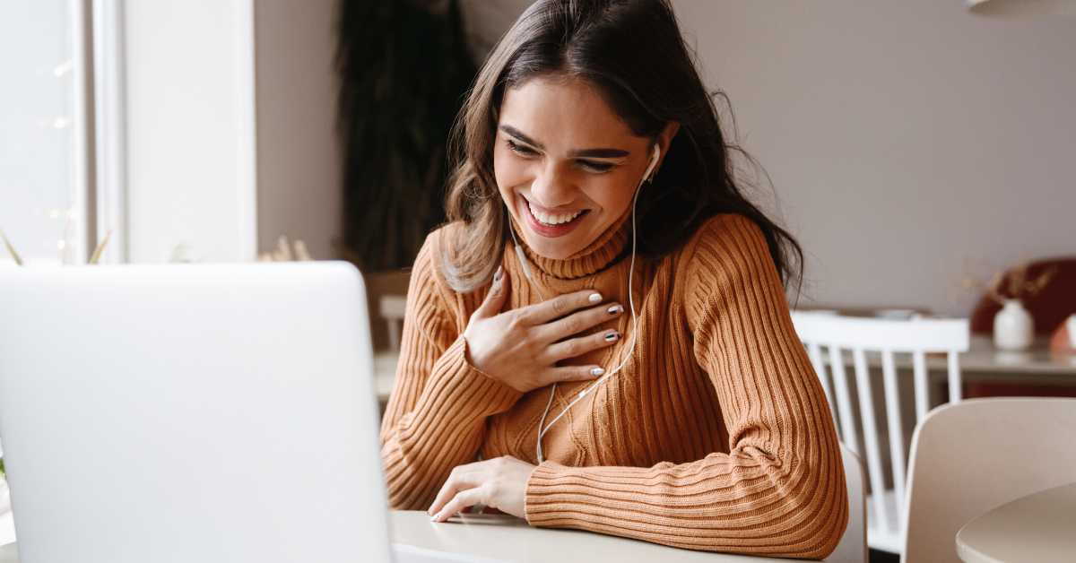A young woman with long dark hair, wearing a rust-colored sweater, is smiling while looking at her laptop. She has earphones in her ears and her hand on her chest. She is in a bright, cozy room with a window and some decorative elements in the background.