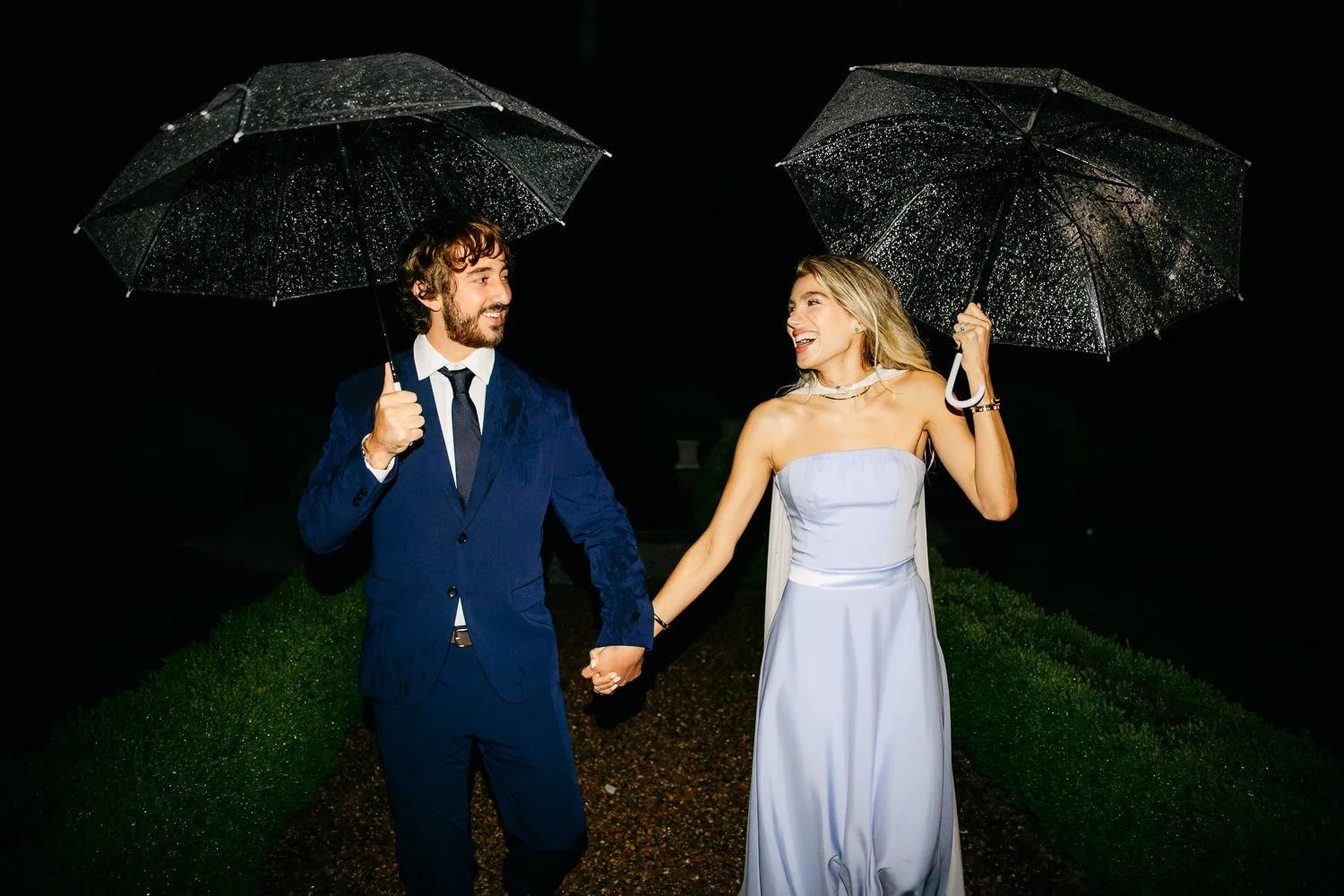 A couple dressed in formal attire, holding umbrellas, walking hand in hand at night, laughing and smiling under the rain.