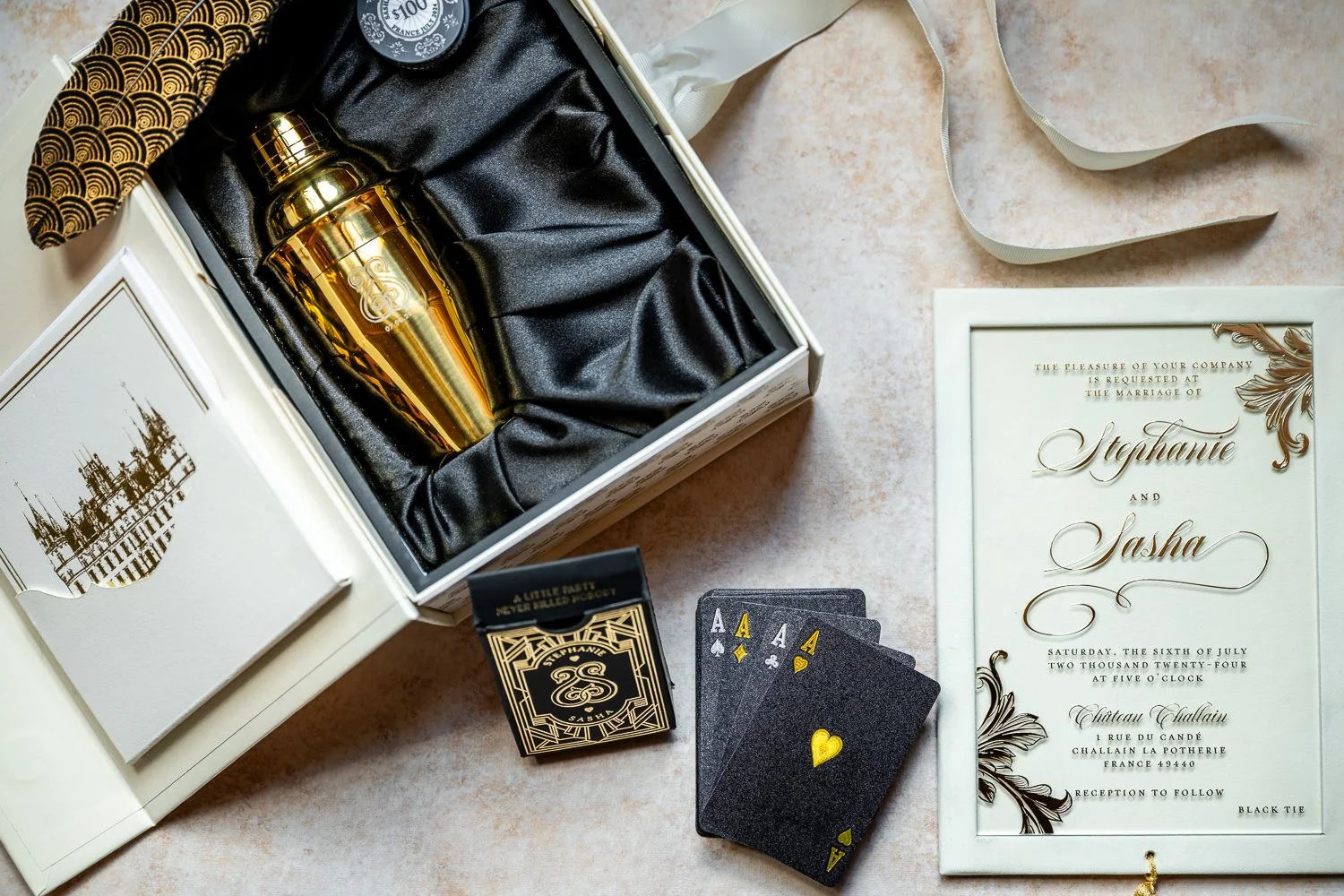 A wedding invitation, a gold cocktail shaker, a deck of playing cards, a box of small chocolates, and a gift box are arranged on a light surface, possibly for a wedding or celebration.