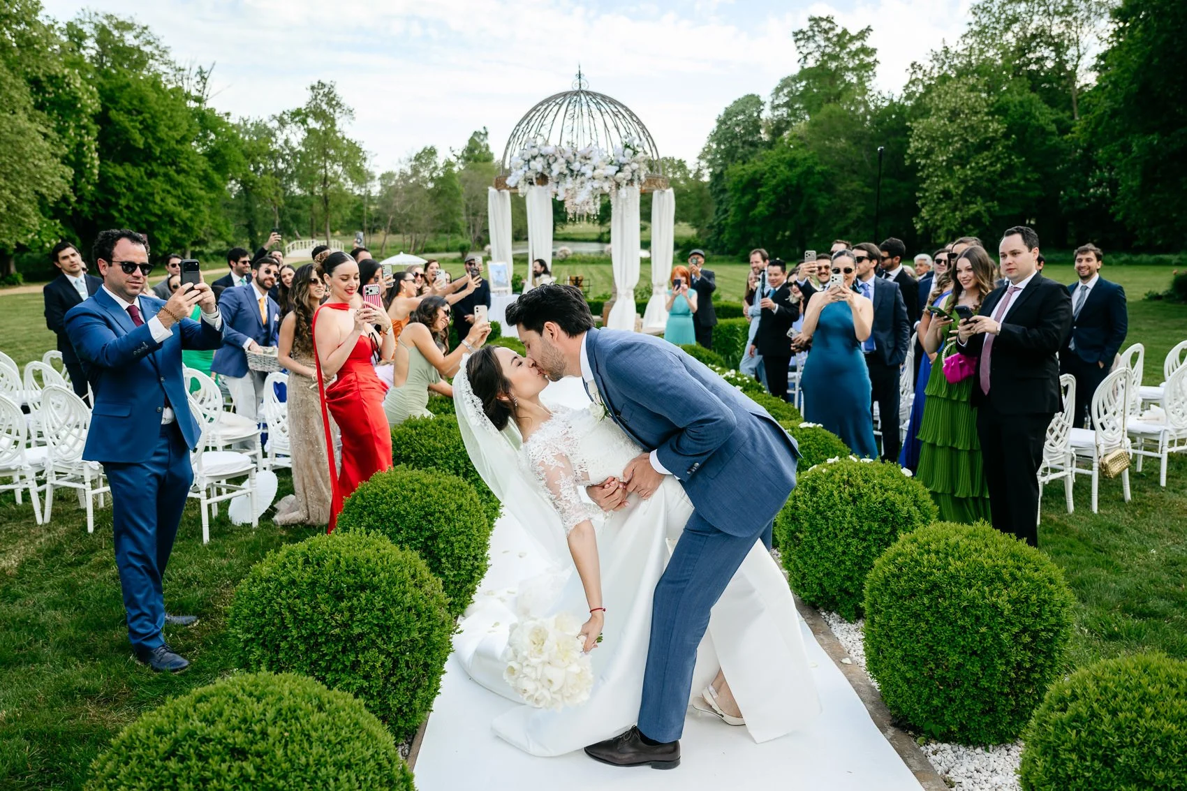 A wedding ceremony outdoors with a bride and groom sharing a kiss, surrounded by guests taking photos in the lush green garden.