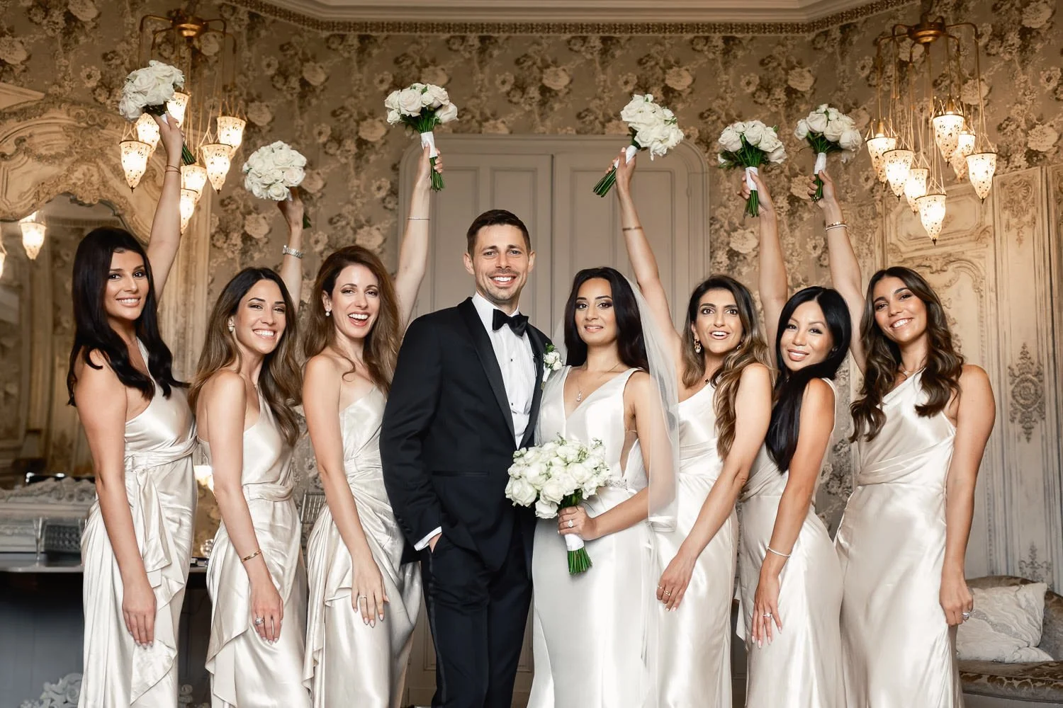 A bride and groom posing with bridesmaids at a wedding. The bride holds a bouquet, and the bridesmaids hold smaller bouquets raised in the air. The group is standing inside a decorated room with ornate wallpaper and chandeliers.