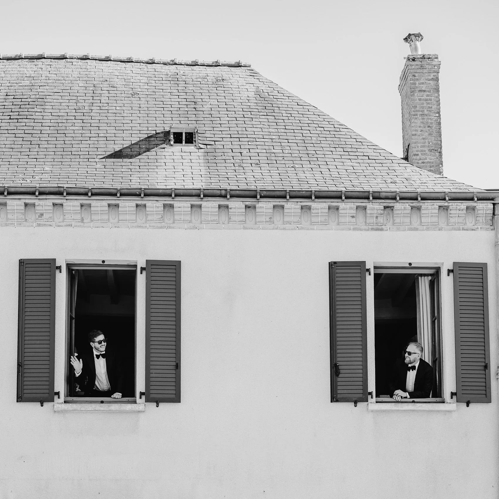 Black and white photo of two men in tuxedos with sunglasses, each standing at open window shutters on a building. One man waves, the other looks out with a smile. The building has a tiled roof and a chimney on the right.