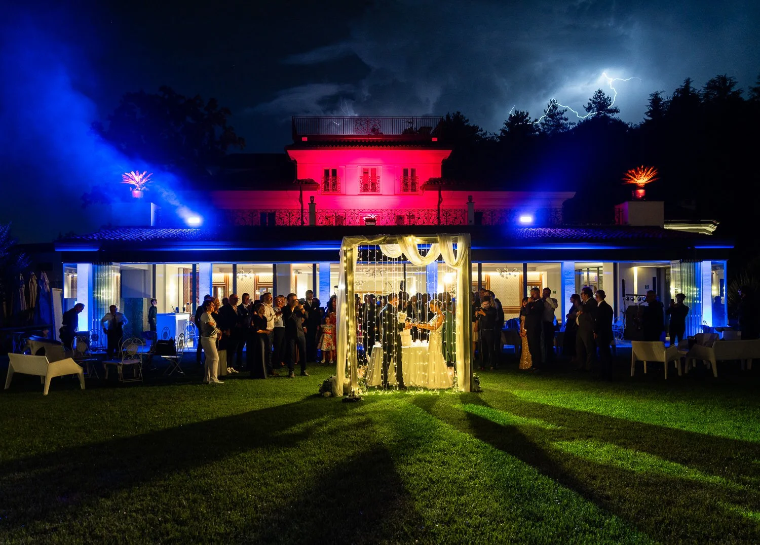 Nighttime wedding reception outdoors at a large house, with a lit archway where a couple is exchanging vows, surrounded by guests and party lighting, with stormy skies and lightning in the background.