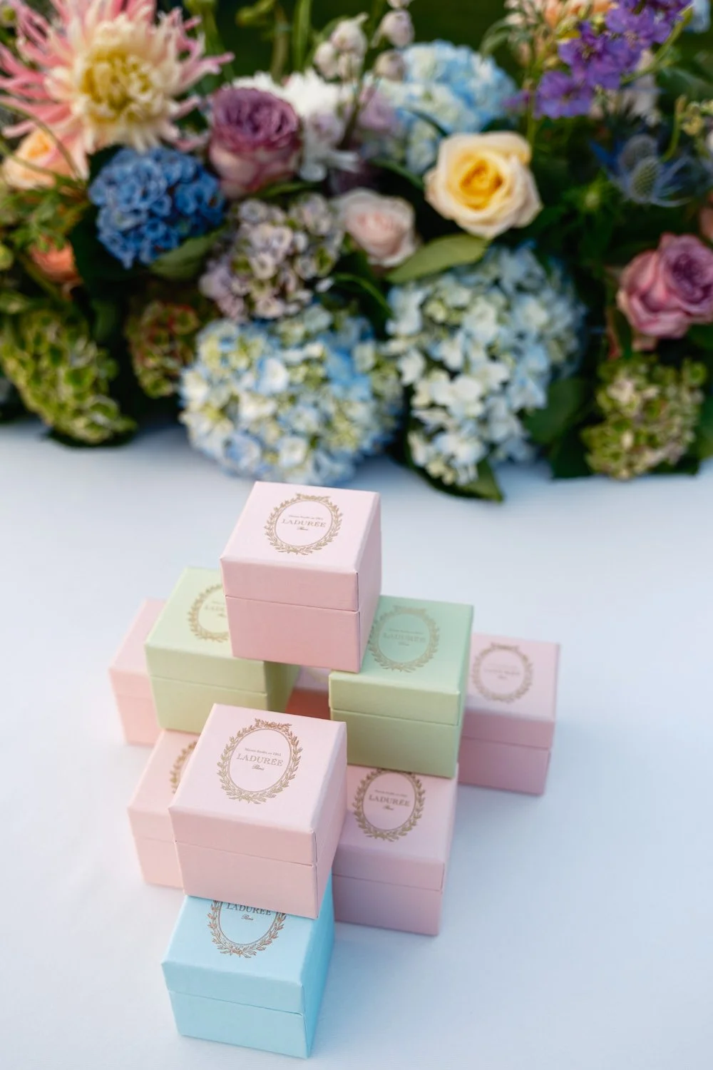 Colorful pastel gift boxes with gold labels on a white surface, with a large bouquet of mixed flowers in the background.
