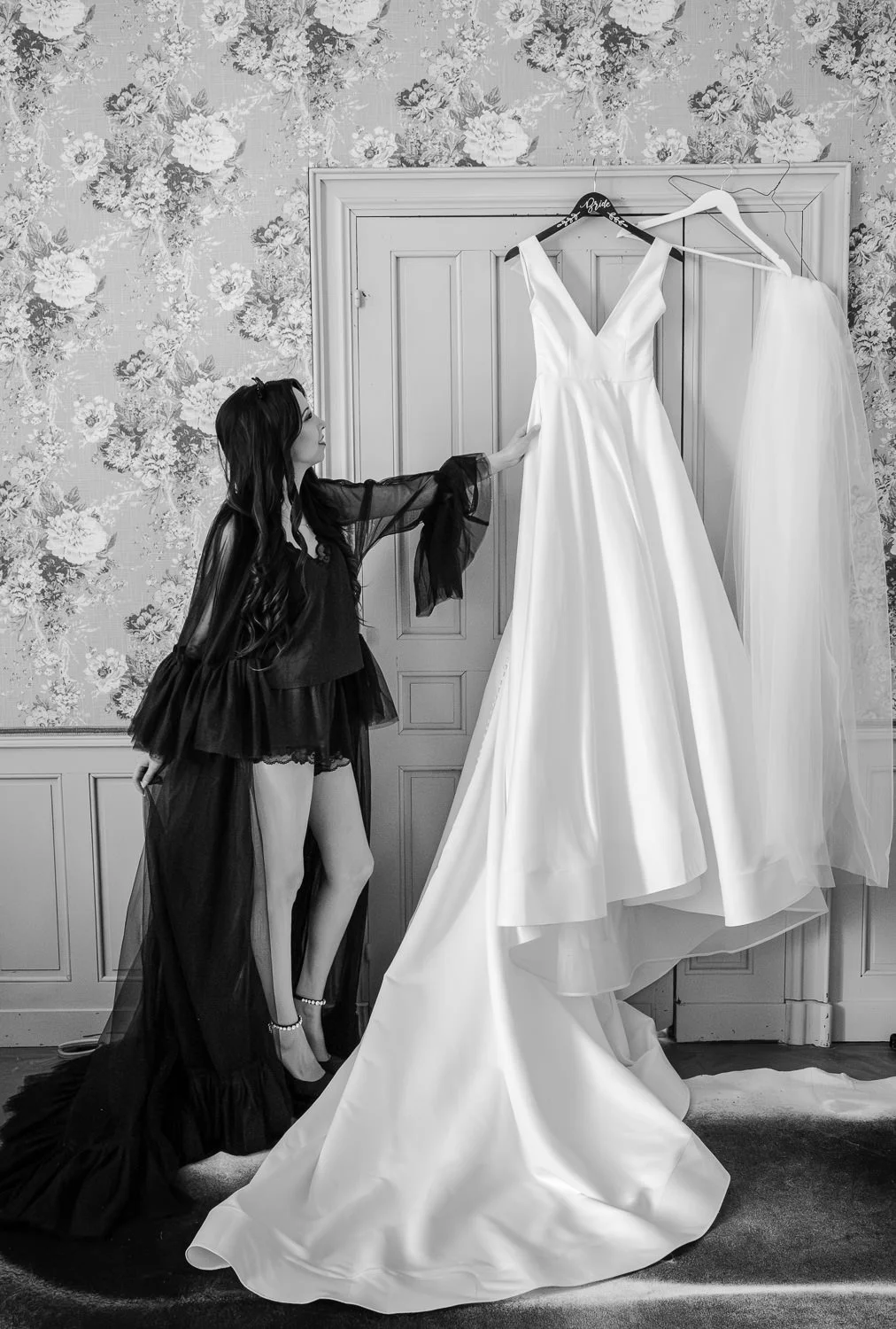 A woman in black lingerie and a sheer robe is reaching out towards a white wedding dress hanging on a door. Next to it is a matching veil. The room has floral wallpaper and decorative paneling.