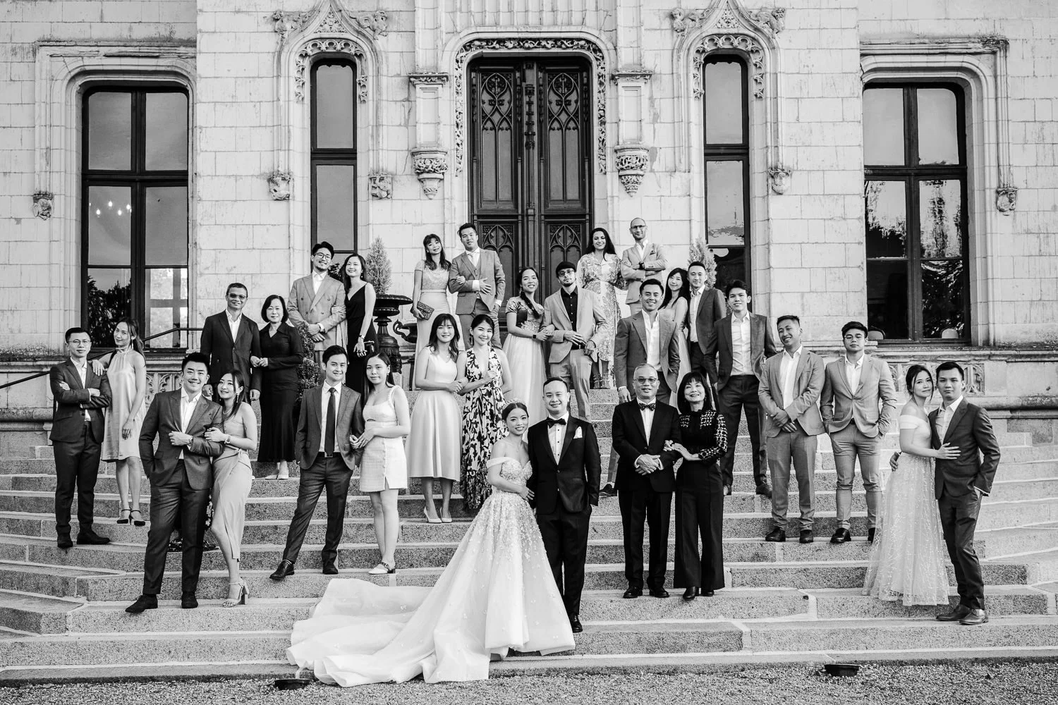 Group of wedding guests on the steps of a historic building, with the bride and groom in the front, black and white photograph.