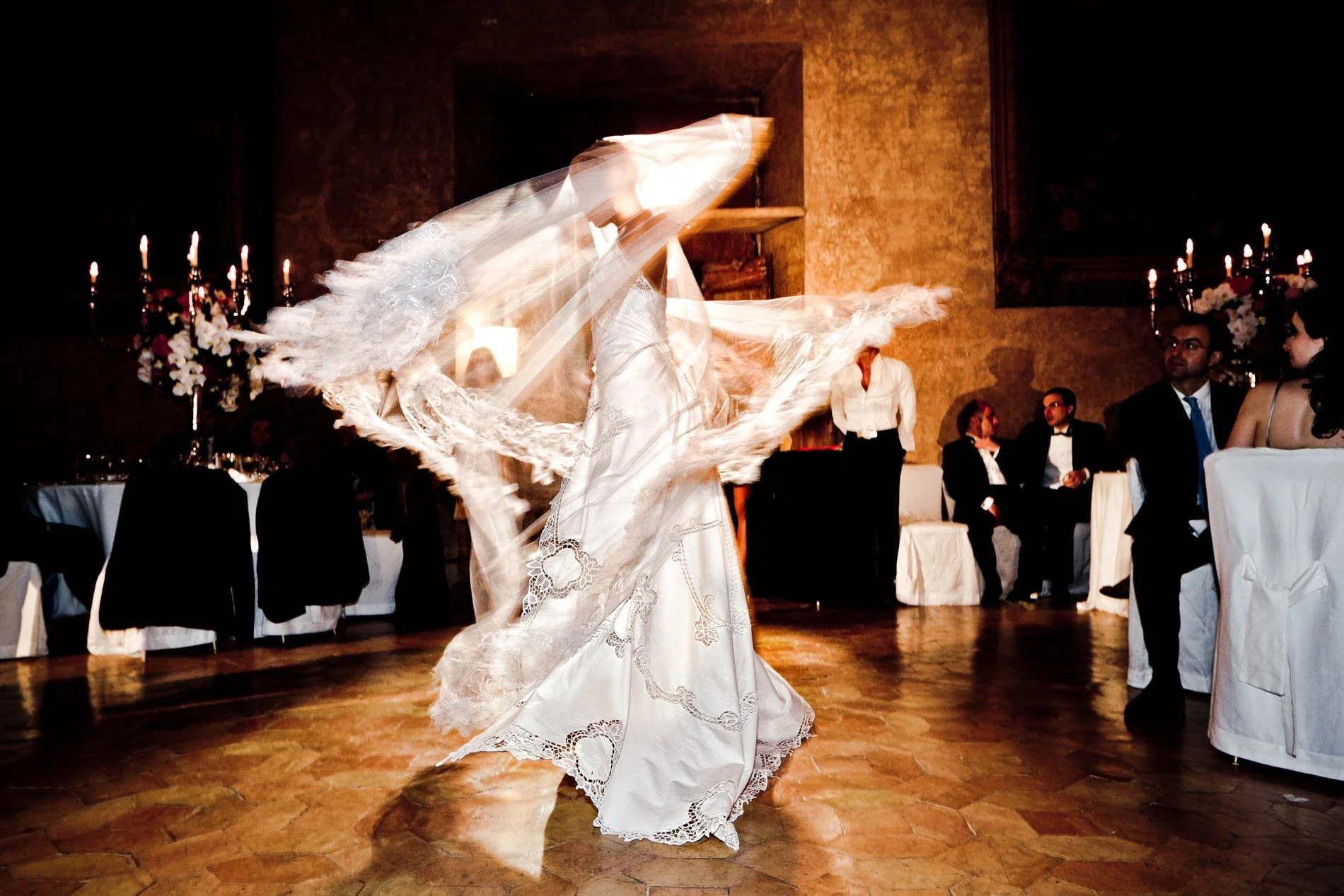 A bride dancing in a white wedding dress with lace details at a wedding reception, with guests seated at banquet tables in the background.