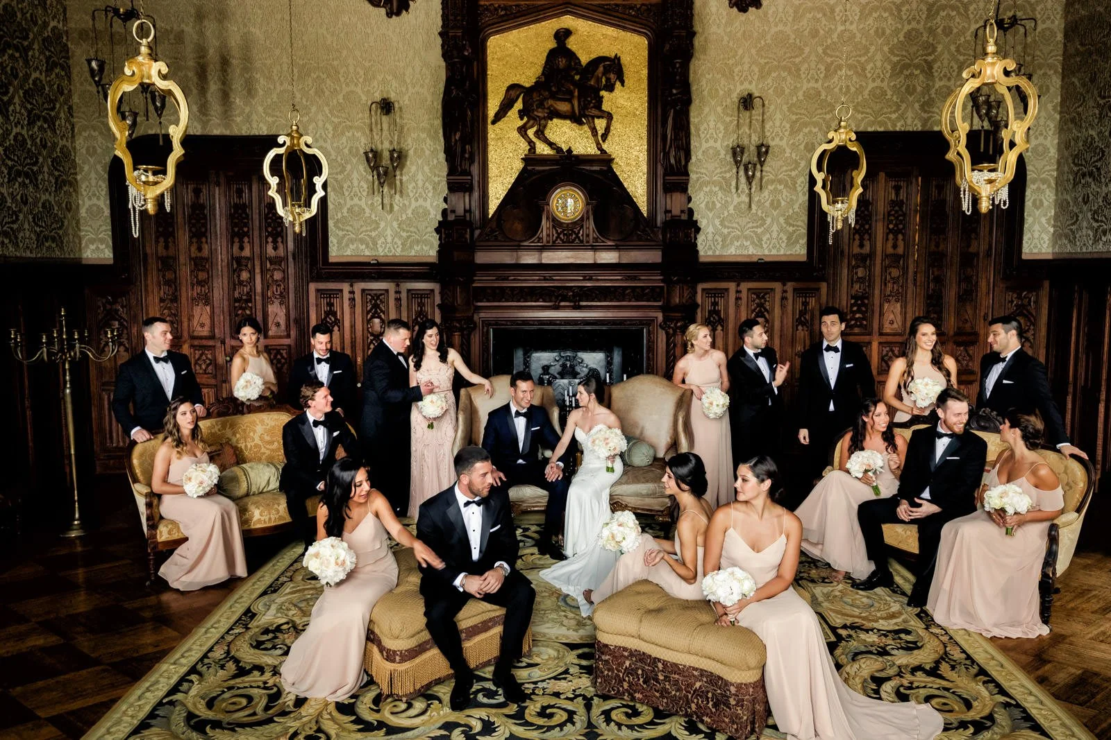 A wedding party in a luxurious, wood-paneled room with ornate décor and gold accents. The group includes men in tuxedos and women in light pink dresses, some holding bouquets, gathered around a fireplace with an equestrian statue above it.