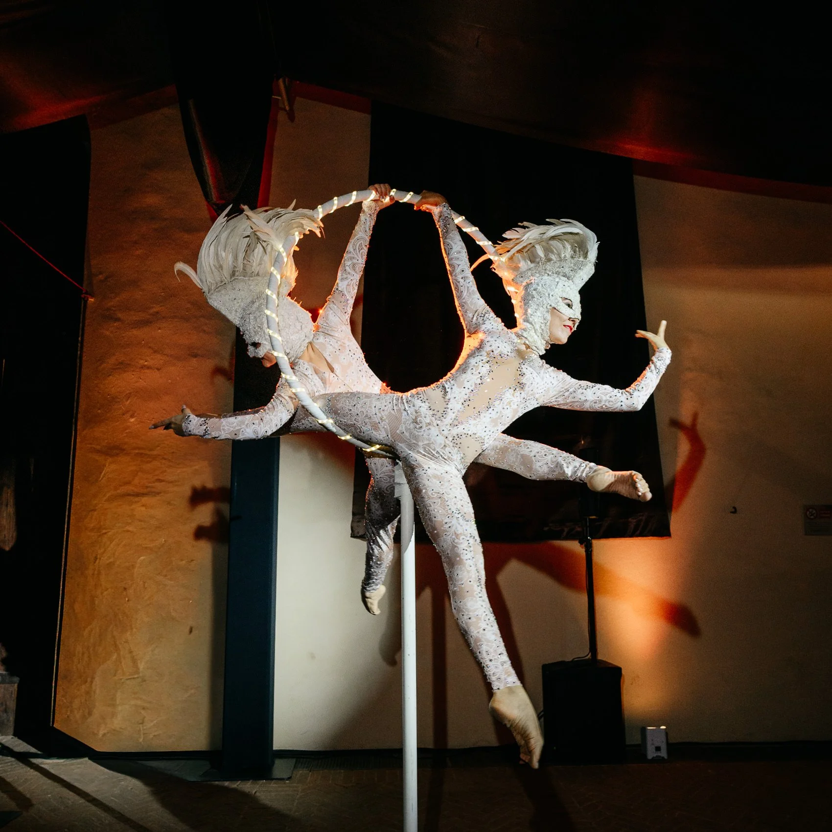 A performer in an elaborate silver costume with feathered headgear poses on a pole in a dimly lit indoor space.