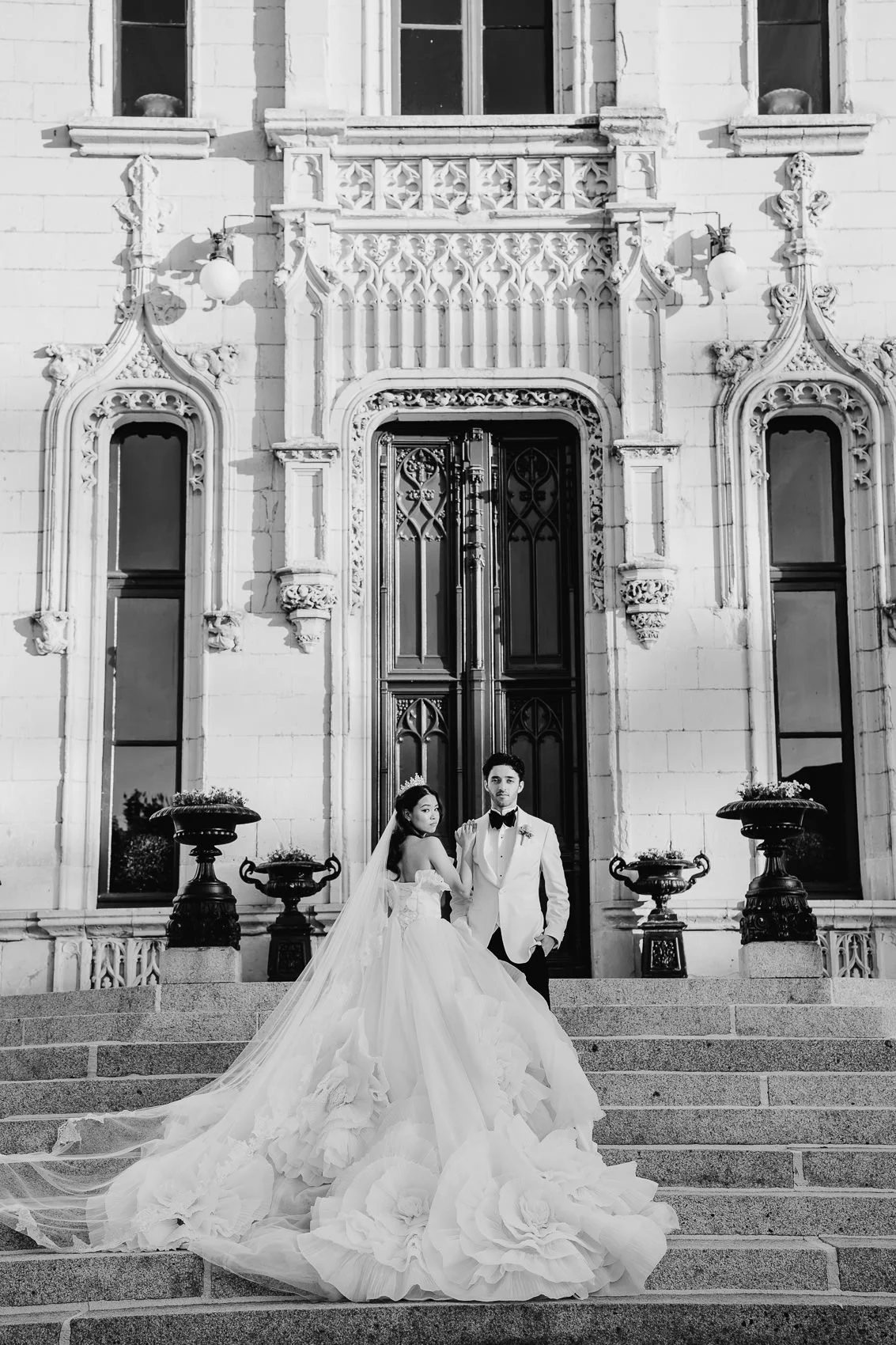 Black and white photo of a bride and groom standing on steps in front of an ornate historic building. The bride wears a voluminous wedding gown with a long train, and the groom is in a white tuxedo with a bow tie. The building features decorative sto