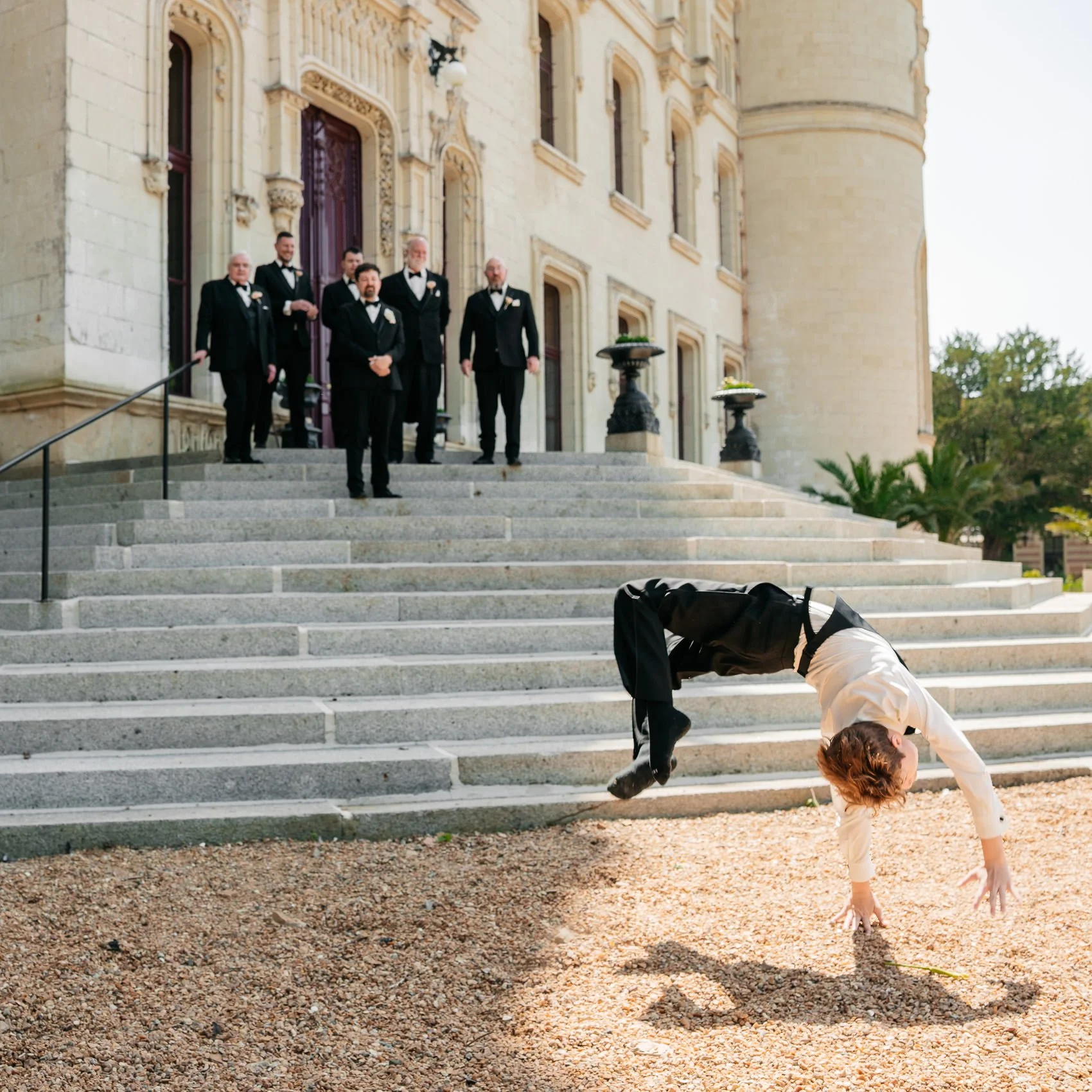 A person in formal attire doing a handstand on steps outside a large, ornate building while a group of men in tuxedos stands on the steps watching.