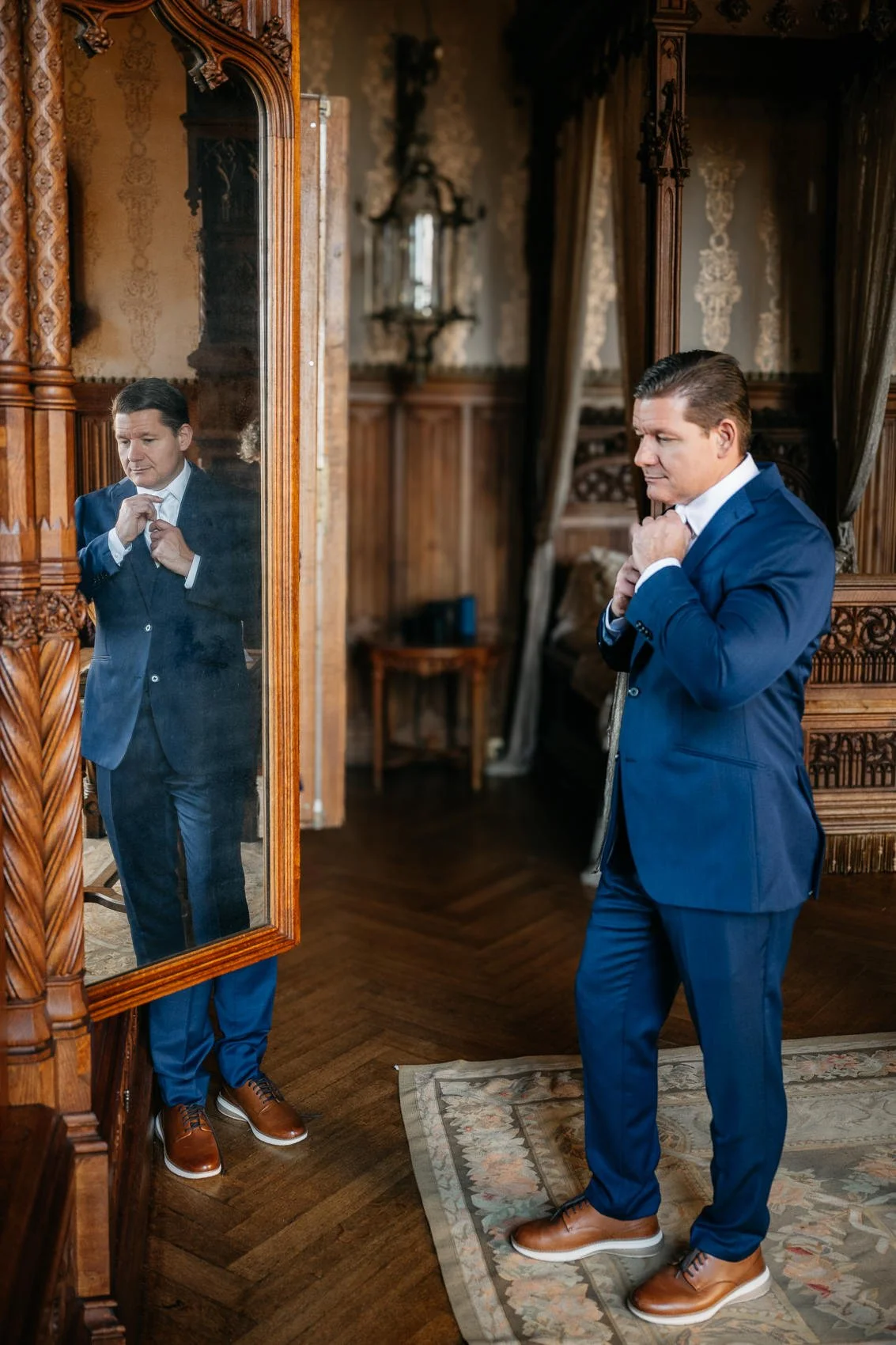 A man in a blue suit adjusting his tie while looking at himself in a tall, ornate mirror in a richly decorated room with wooden paneling and patterned walls.