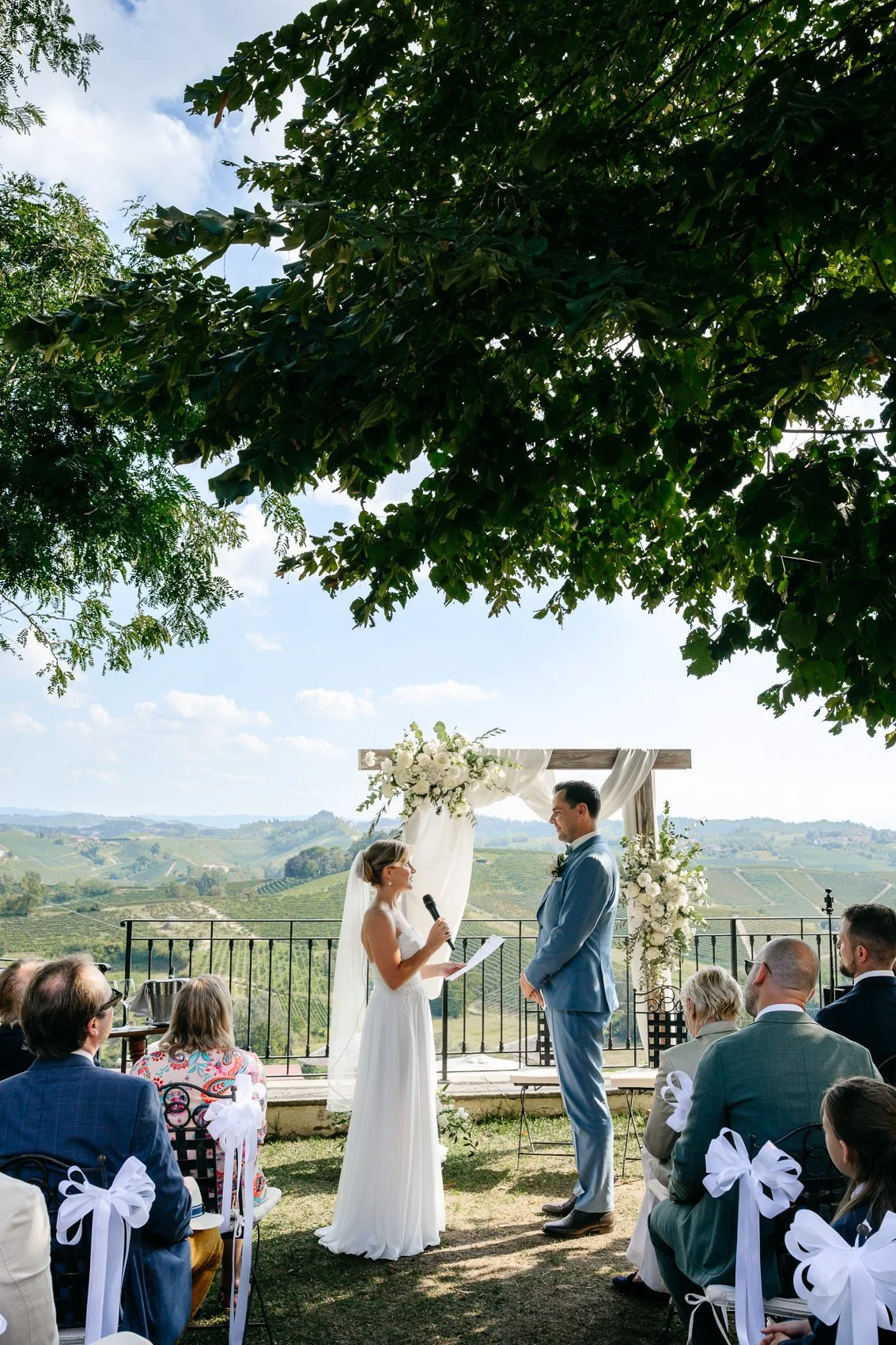 Bride and groom exchanging vows during an outdoor wedding ceremony with a scenic vineyard background and guests seated in front.