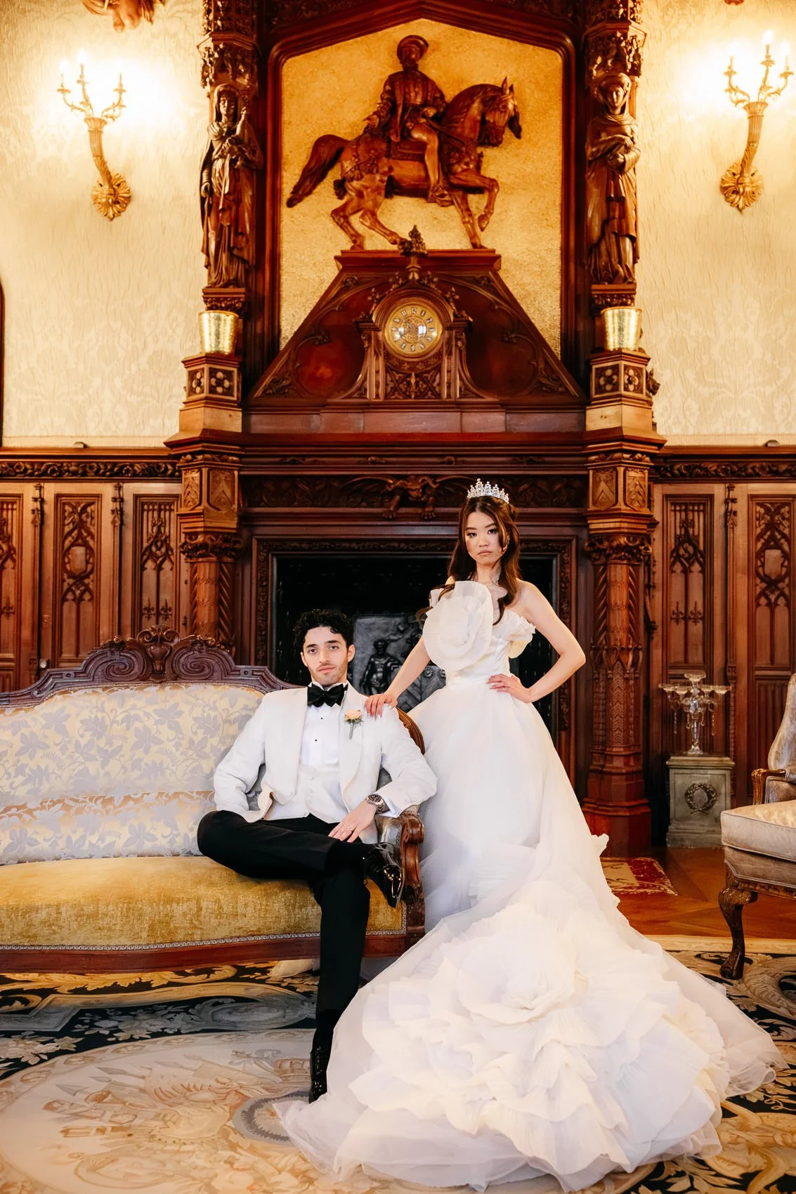 A bride in a white wedding gown with puffed sleeves and a tiara, standing with her hand on a seated groom in a tuxedo, inside an ornate, wood-paneled room with a fireplace and detailed carvings.